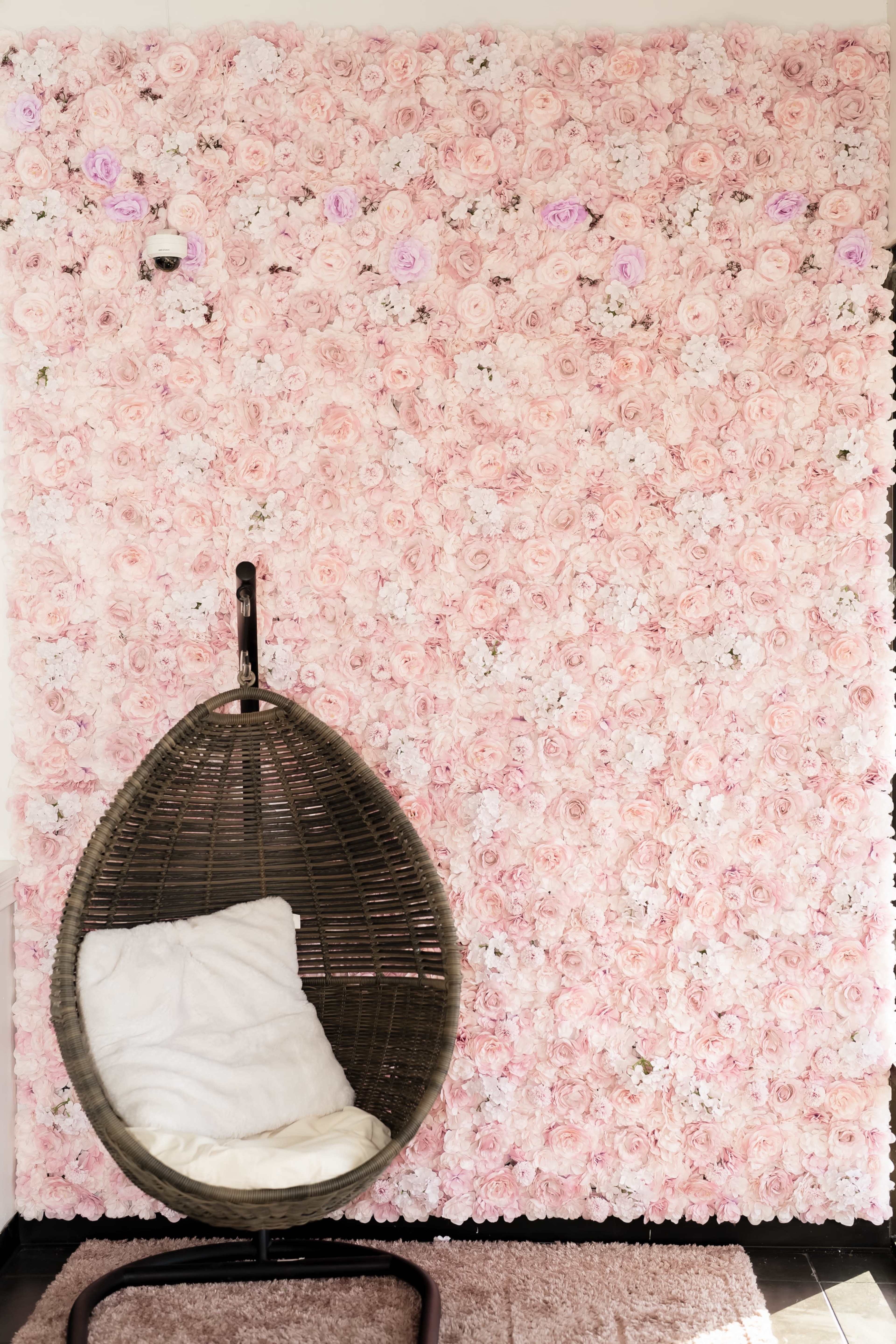 A rattan hanging chair sits in front of a wall covered in pink flowers.