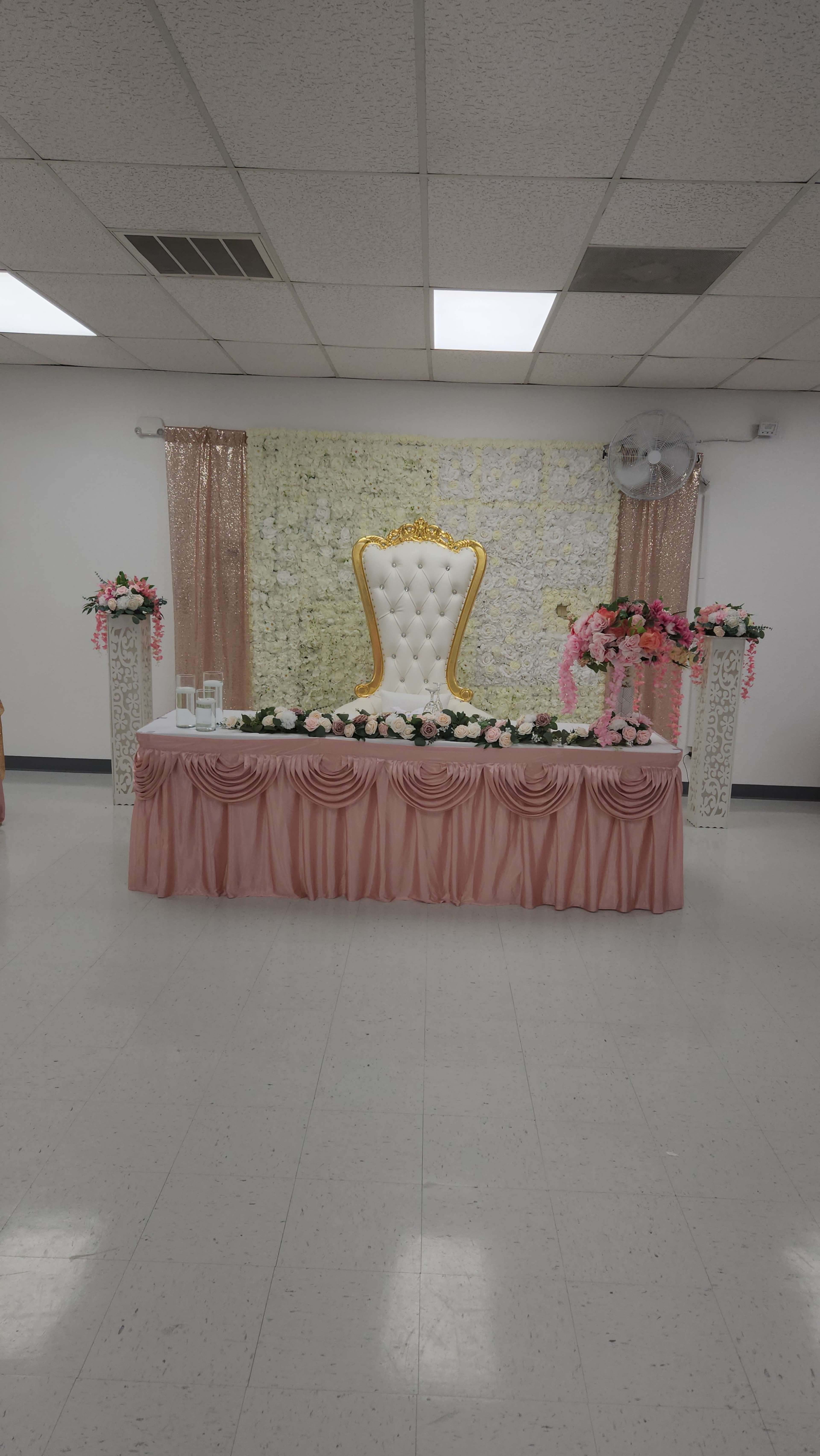 A decorated event space featuring a white throne chair with gold accents, a table draped in pink fabric, and floral arrangements on either side.