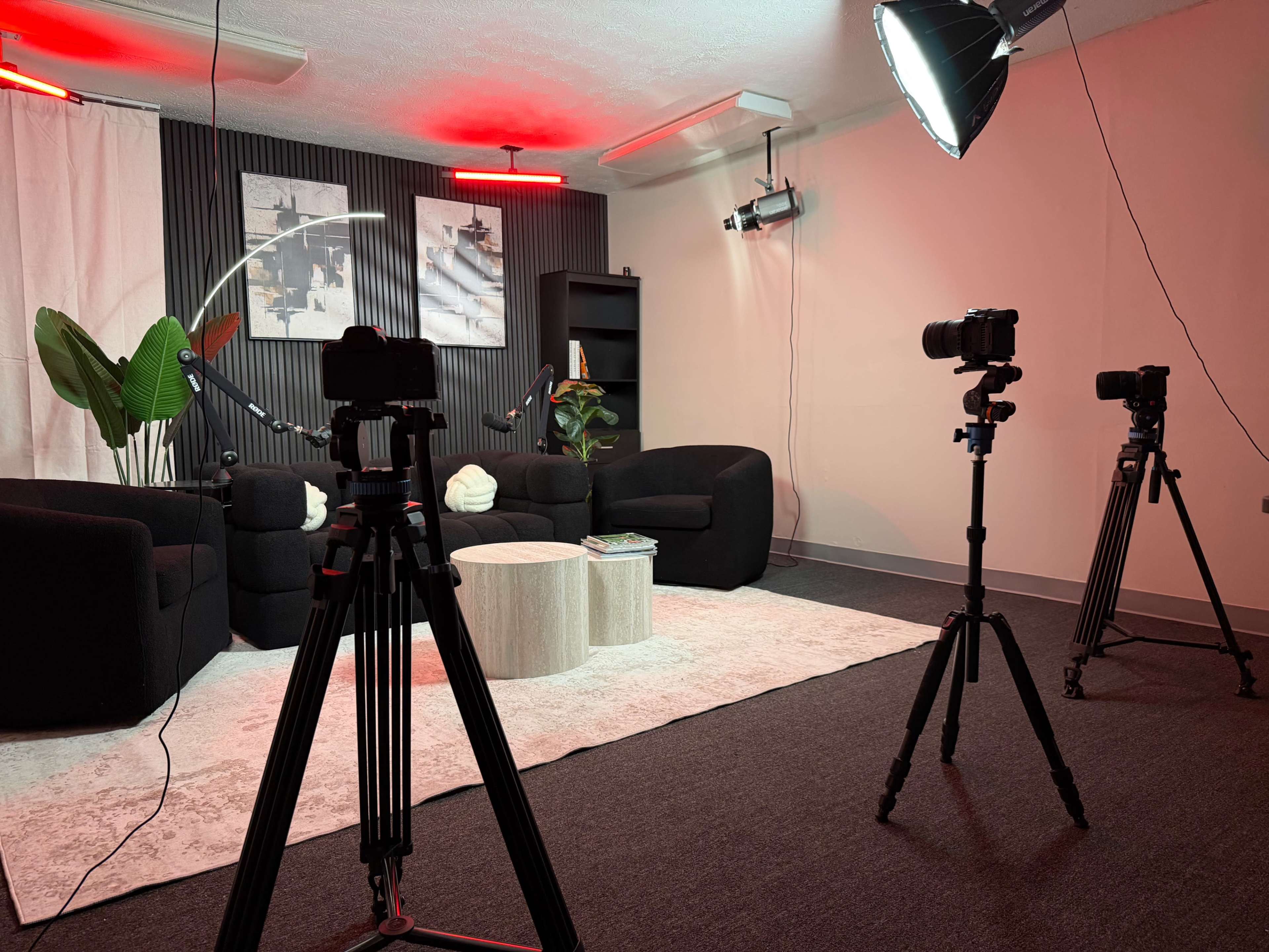 The image shows a modern studio setup with two cameras on tripods facing a seating area that includes two black sofas and a round coffee table, illuminated by red and white lighting.