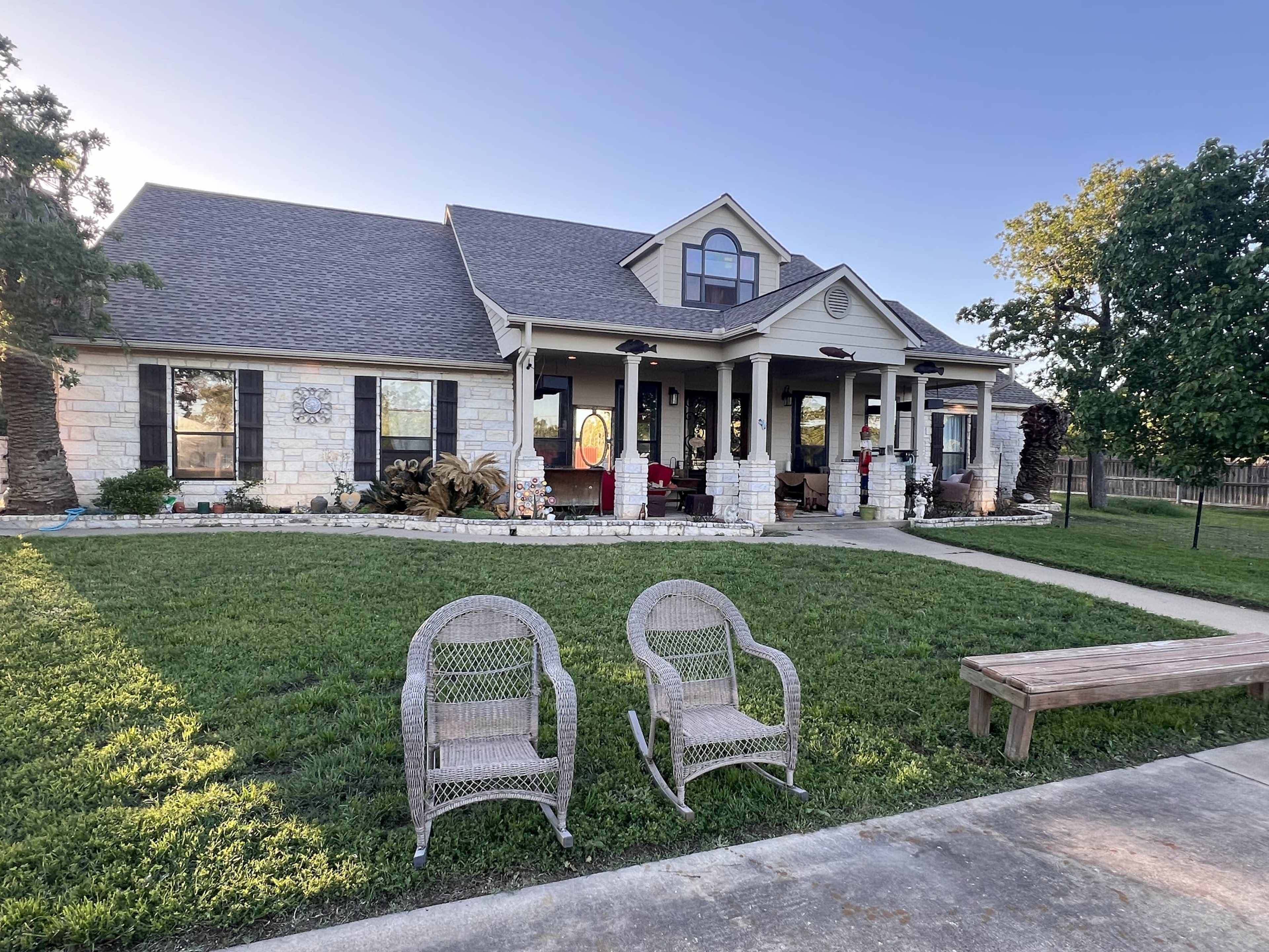 A single-story house with a stone facade features a large front porch, two rocking chairs, and a well-maintained lawn.