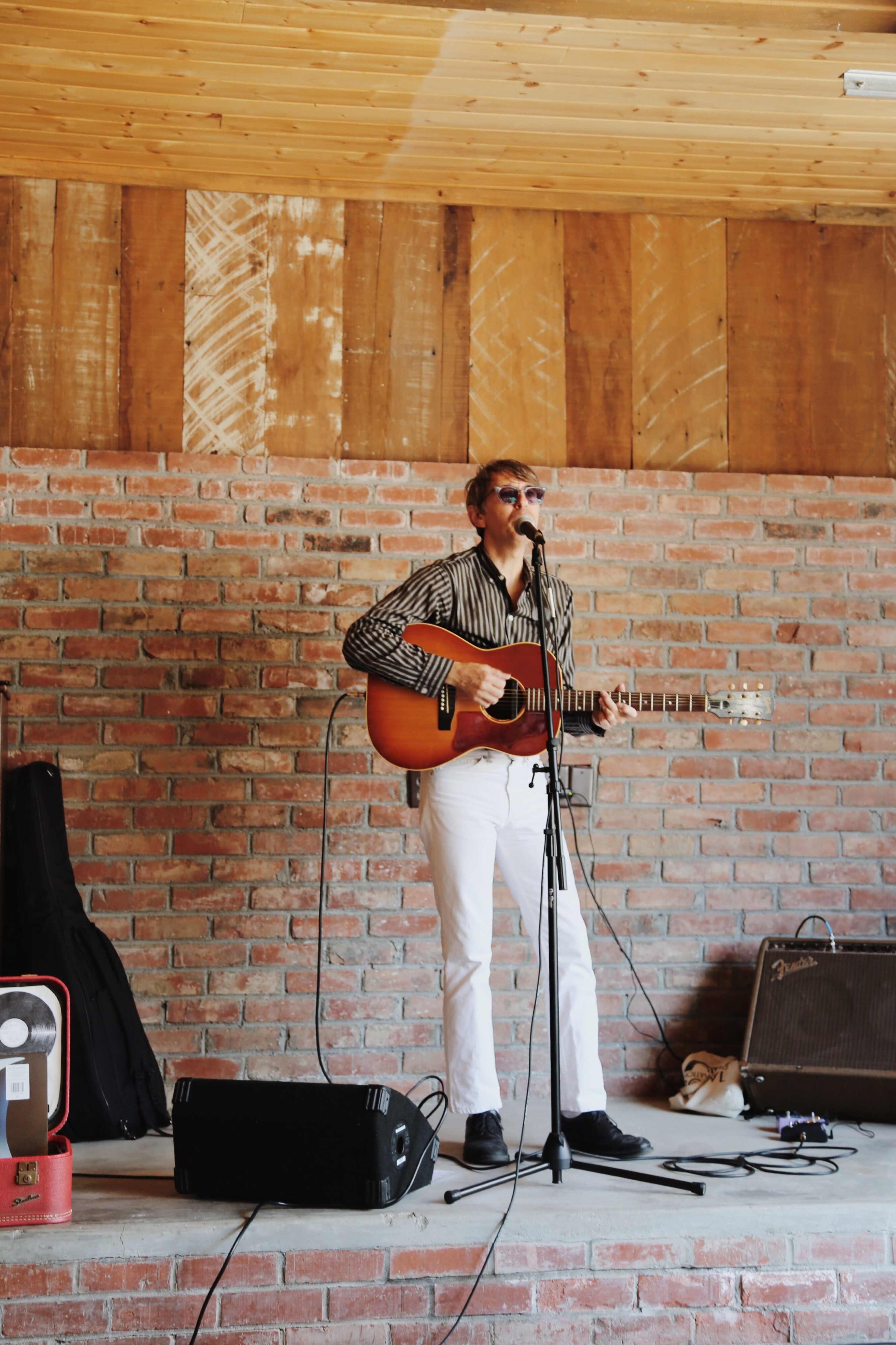 A musician performs on stage with an acoustic guitar in a rustic brick building.