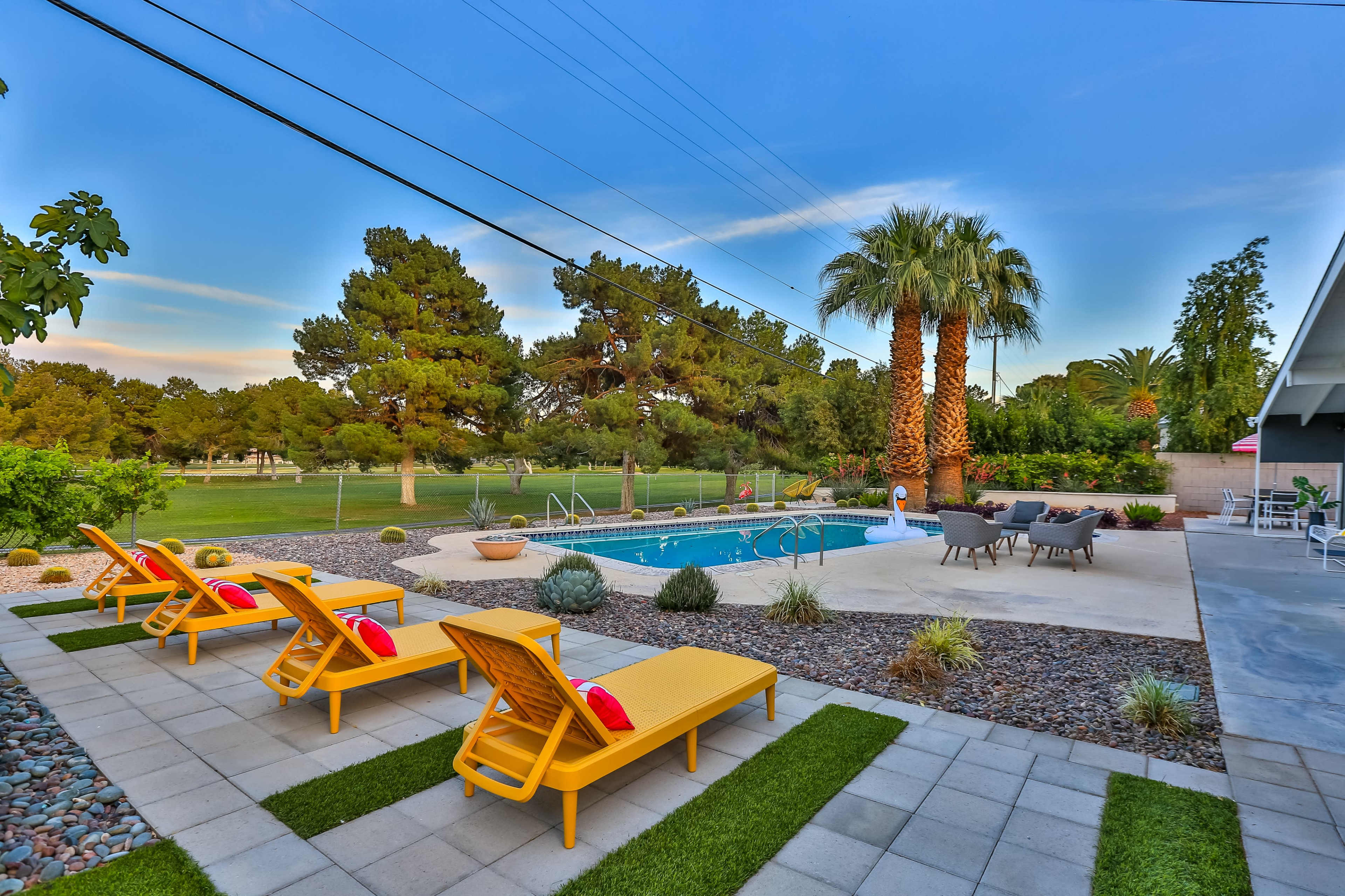 A backyard features a swimming pool surrounded by lounge chairs, palm trees, and landscaped greenery.