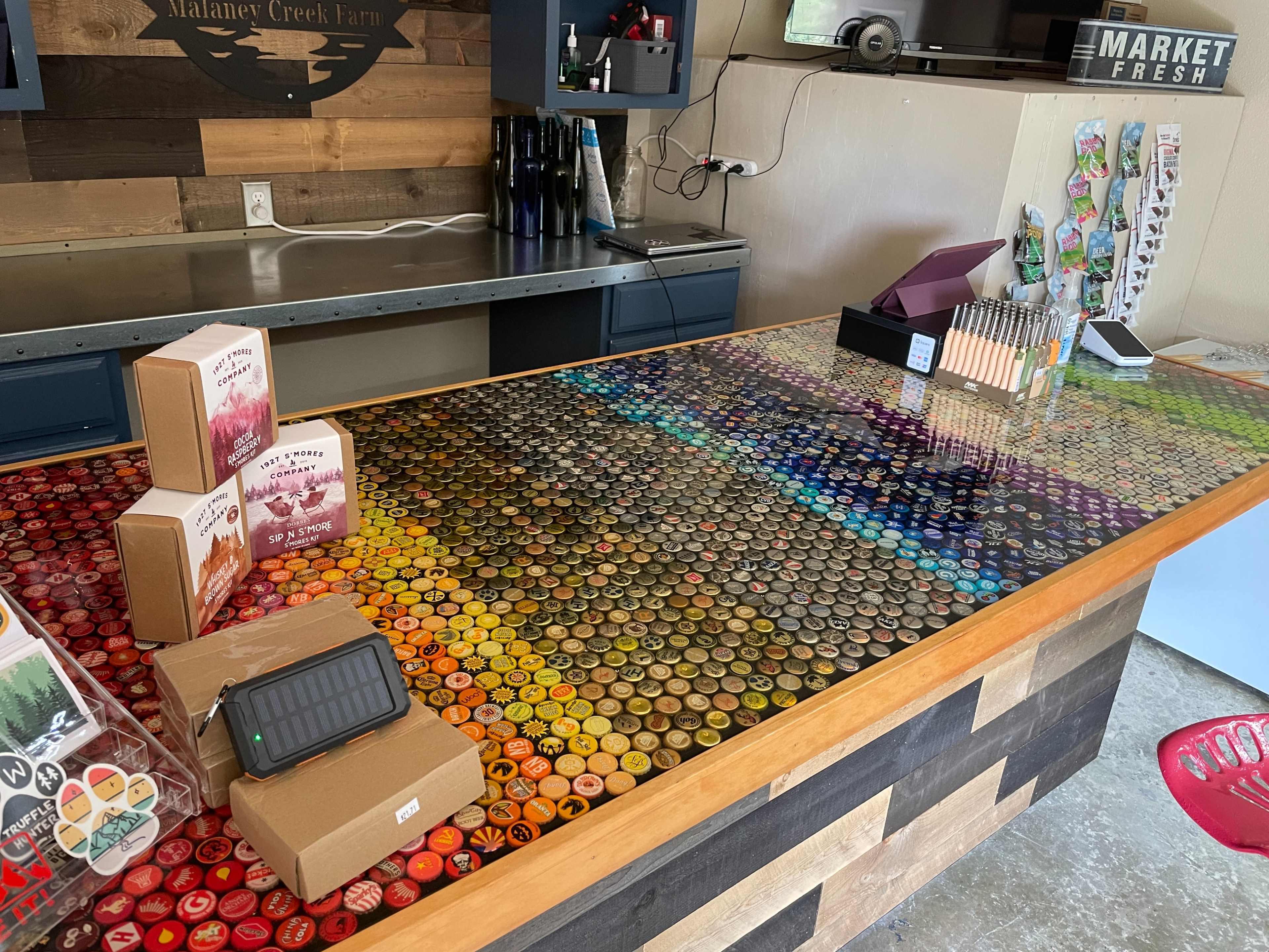A countertop covered with a vibrant array of bottle caps, with boxes and items placed on it in a rustic farm store.