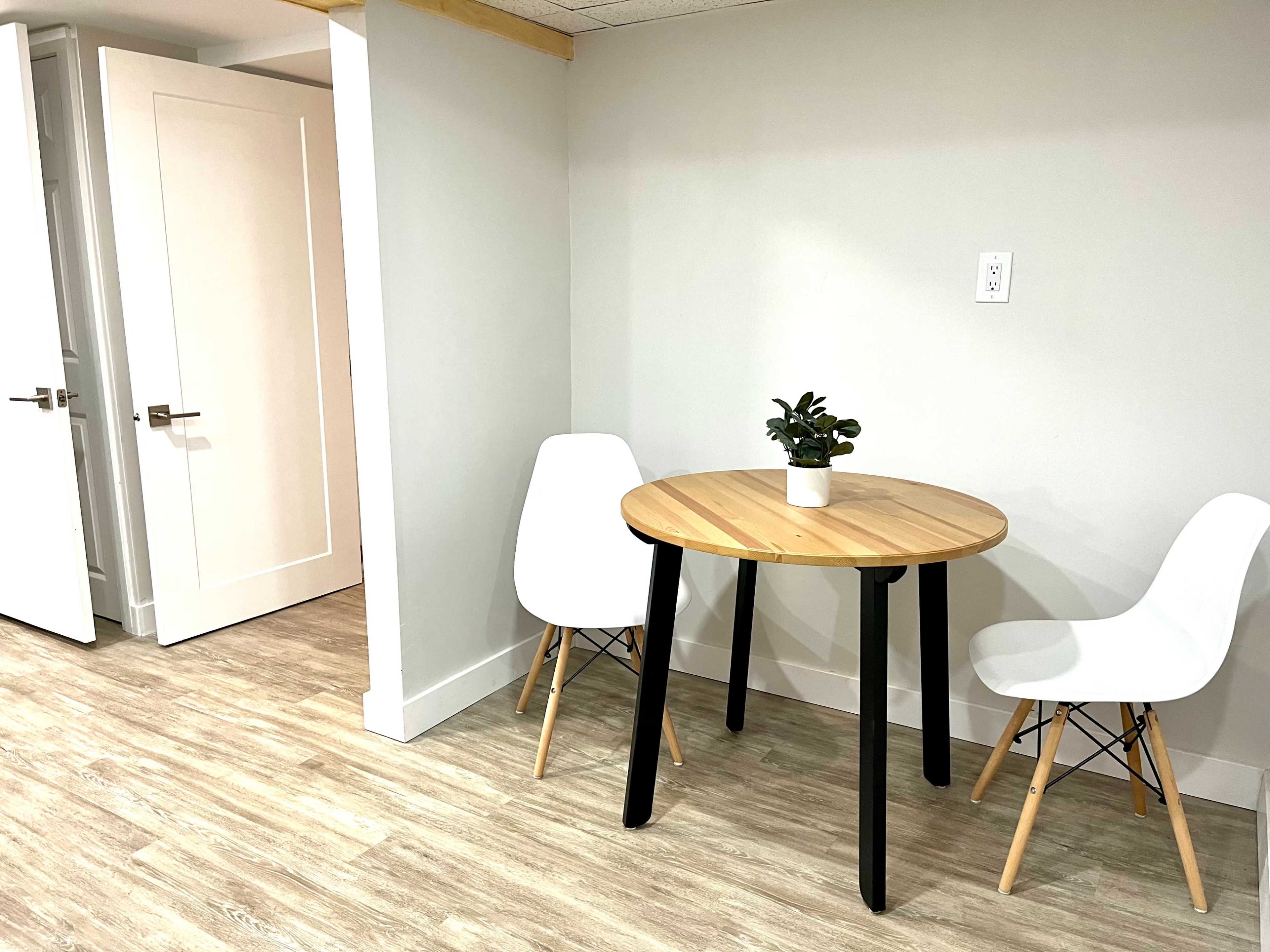 A small dining area features a round wooden table with two white chairs and a potted plant, adjacent to a doorway.