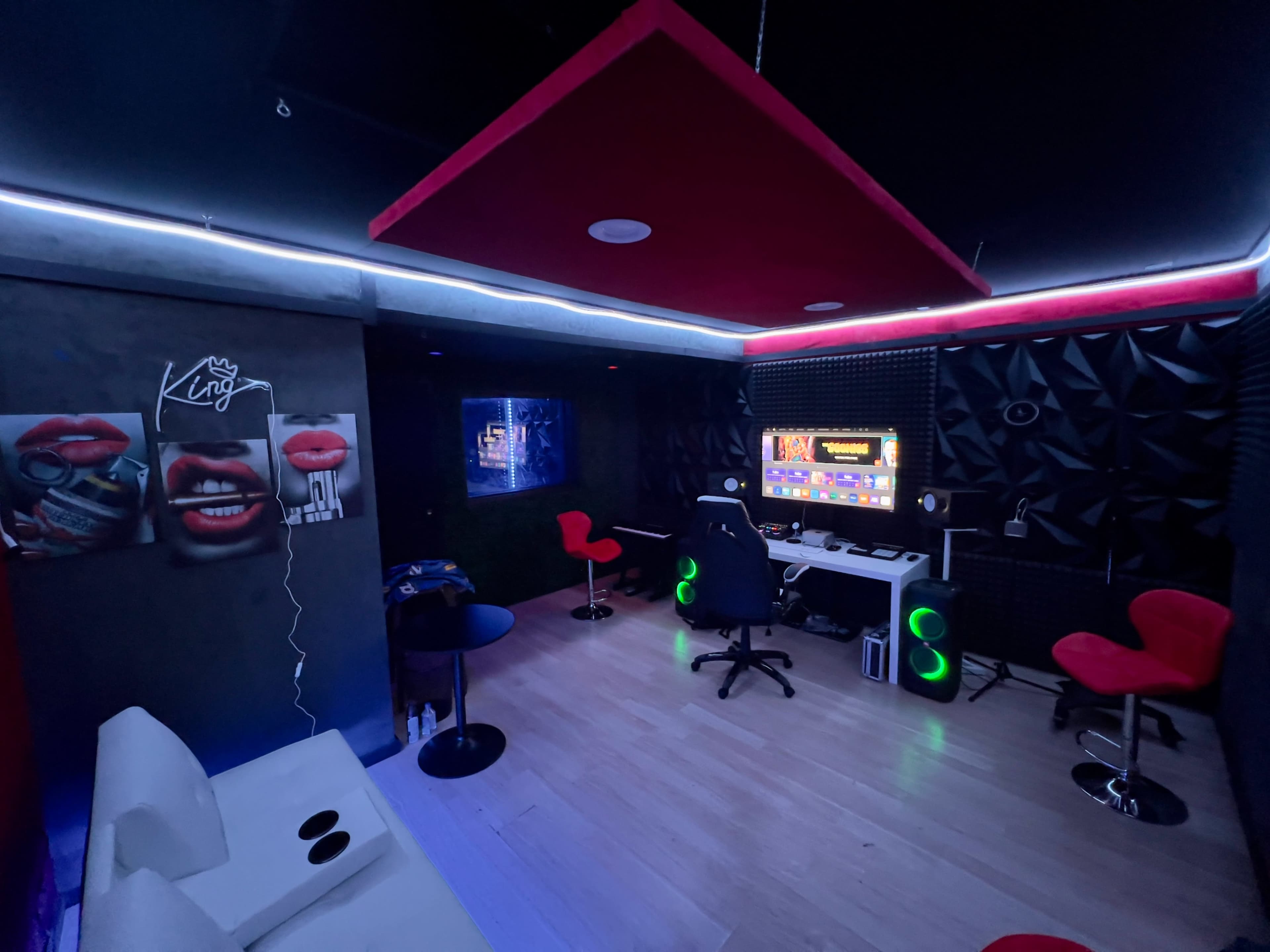 A music studio or recording space with modern equipment, featuring red and green lighting, a white sofa, and two black chairs near a soundproof wall.
