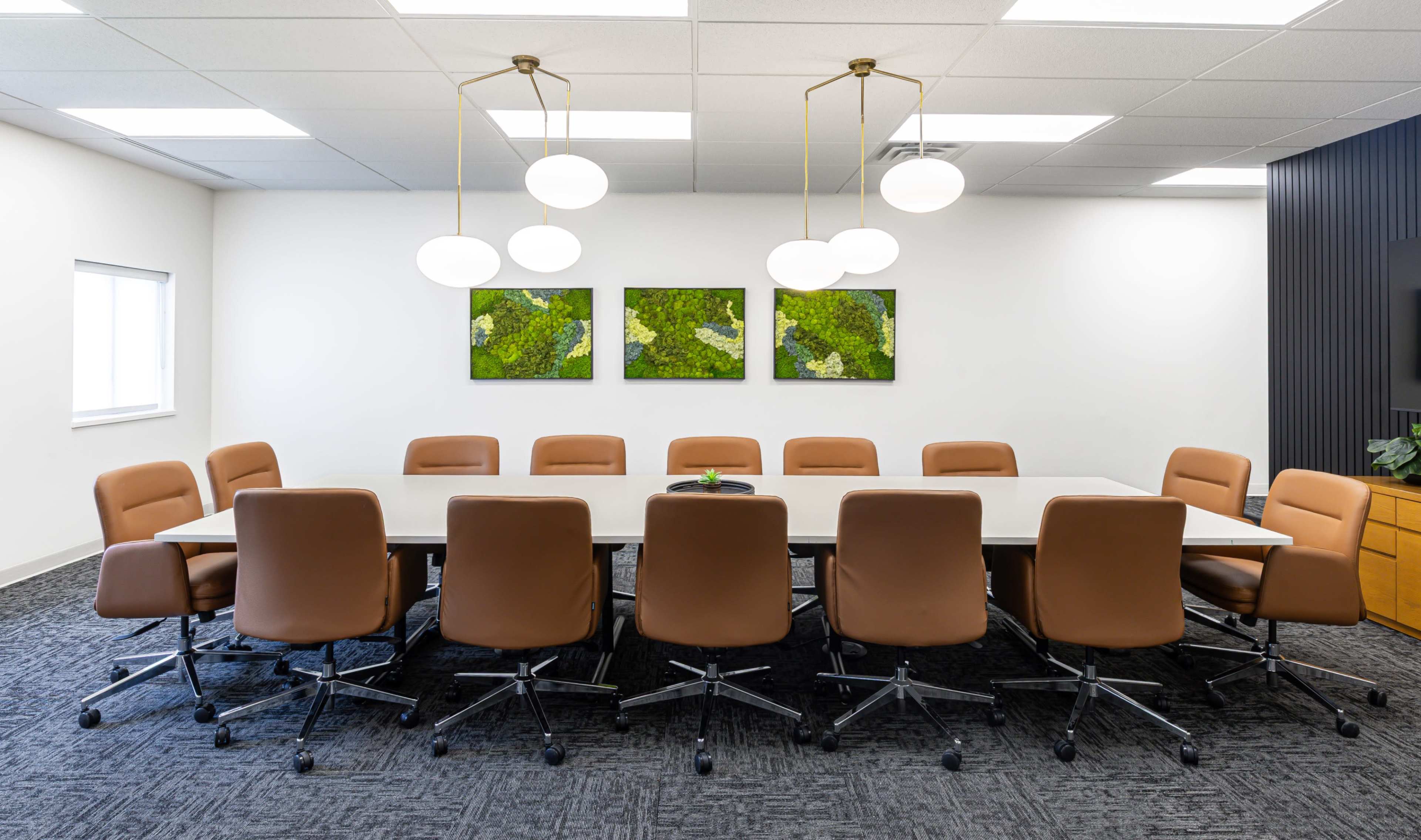 A long conference table surrounded by brown swivel chairs is situated in a well-lit meeting room with three green abstract paintings on the wall.