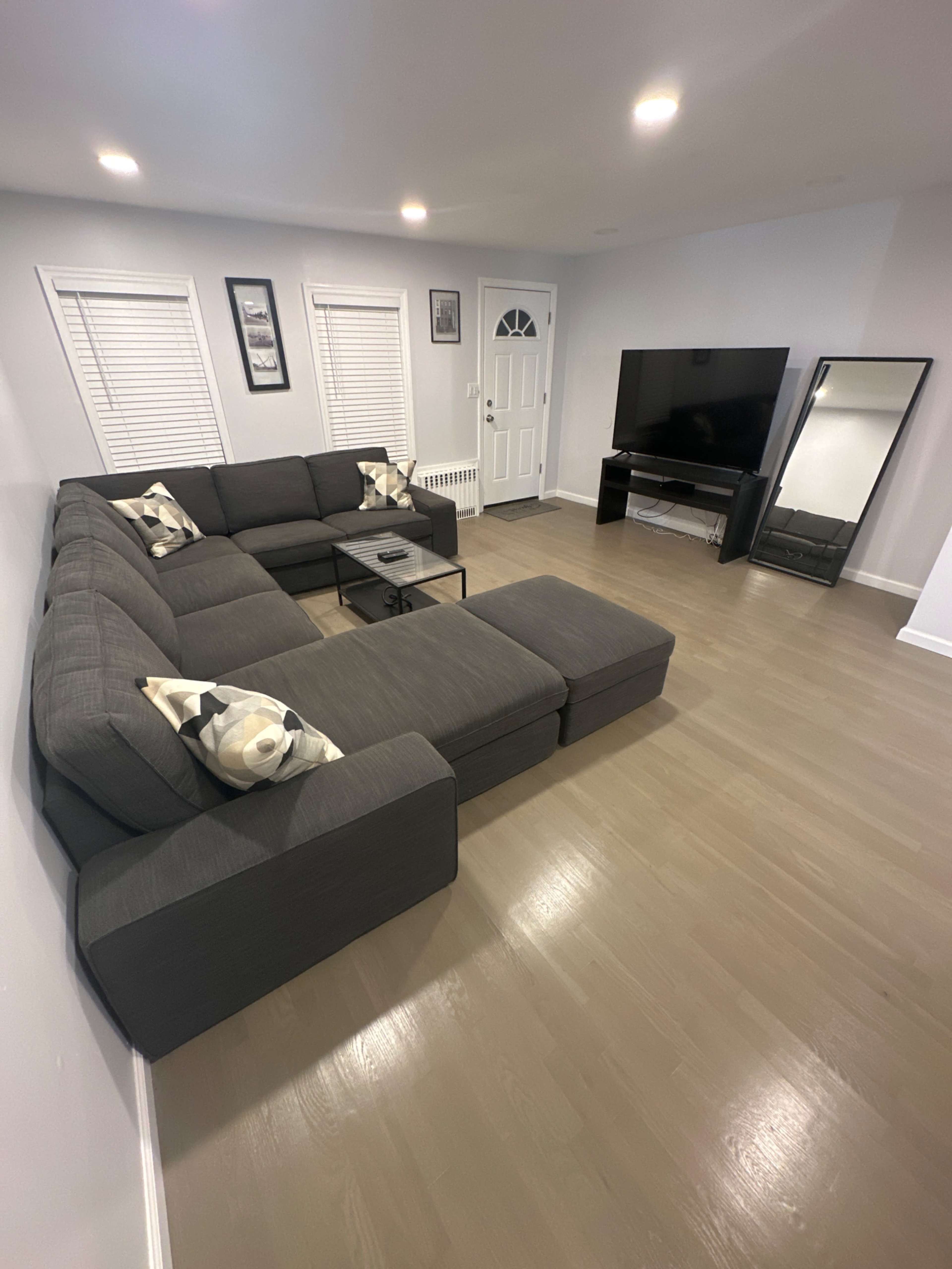 The image shows a modern living room featuring a large sectional sofa, a coffee table, a flat-screen TV, and a full-length mirror.