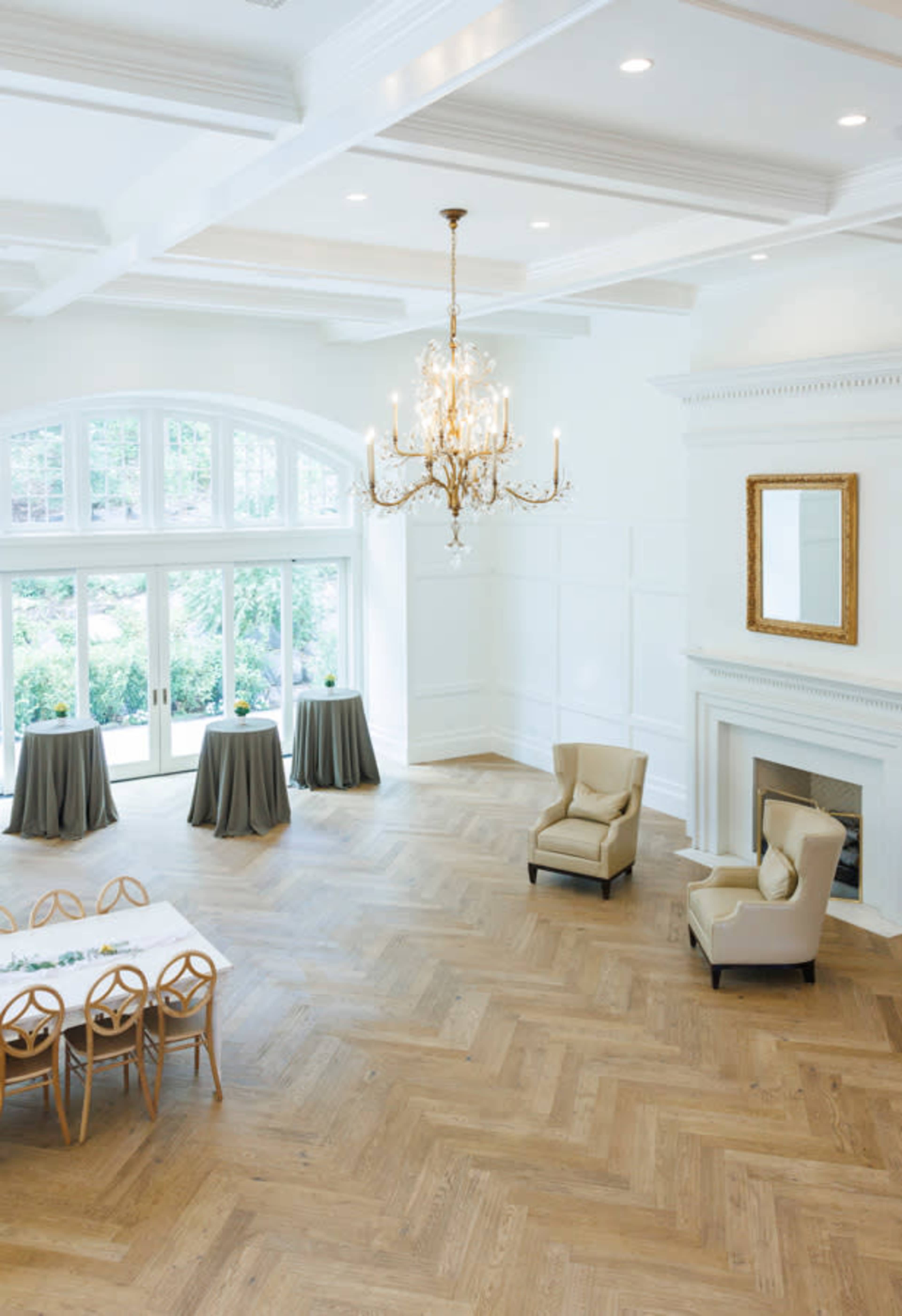 The image shows a spacious, elegantly designed room featuring a chandelier, light-colored walls, and a wooden floor with a seating area and tables set up.