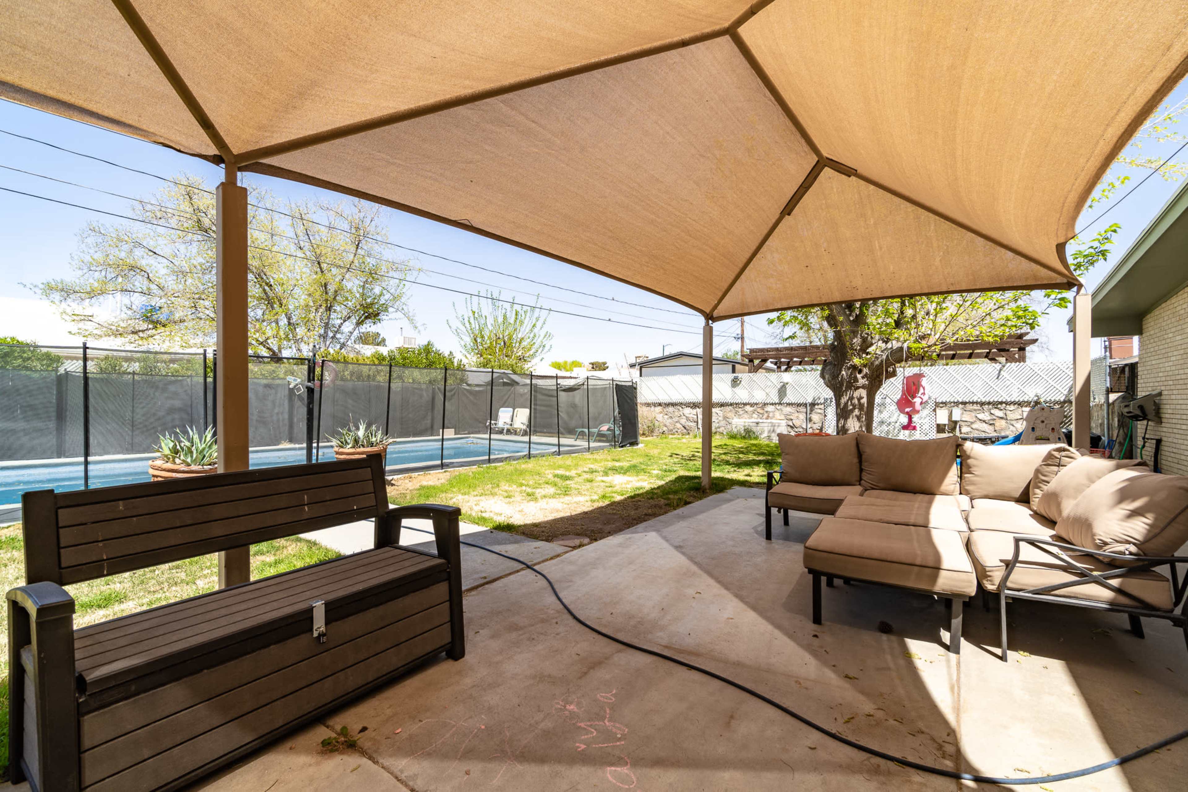 A shaded outdoor space features a seating area with a sectional sofa and a bench, overlooking a pool surrounded by a fence and trees.