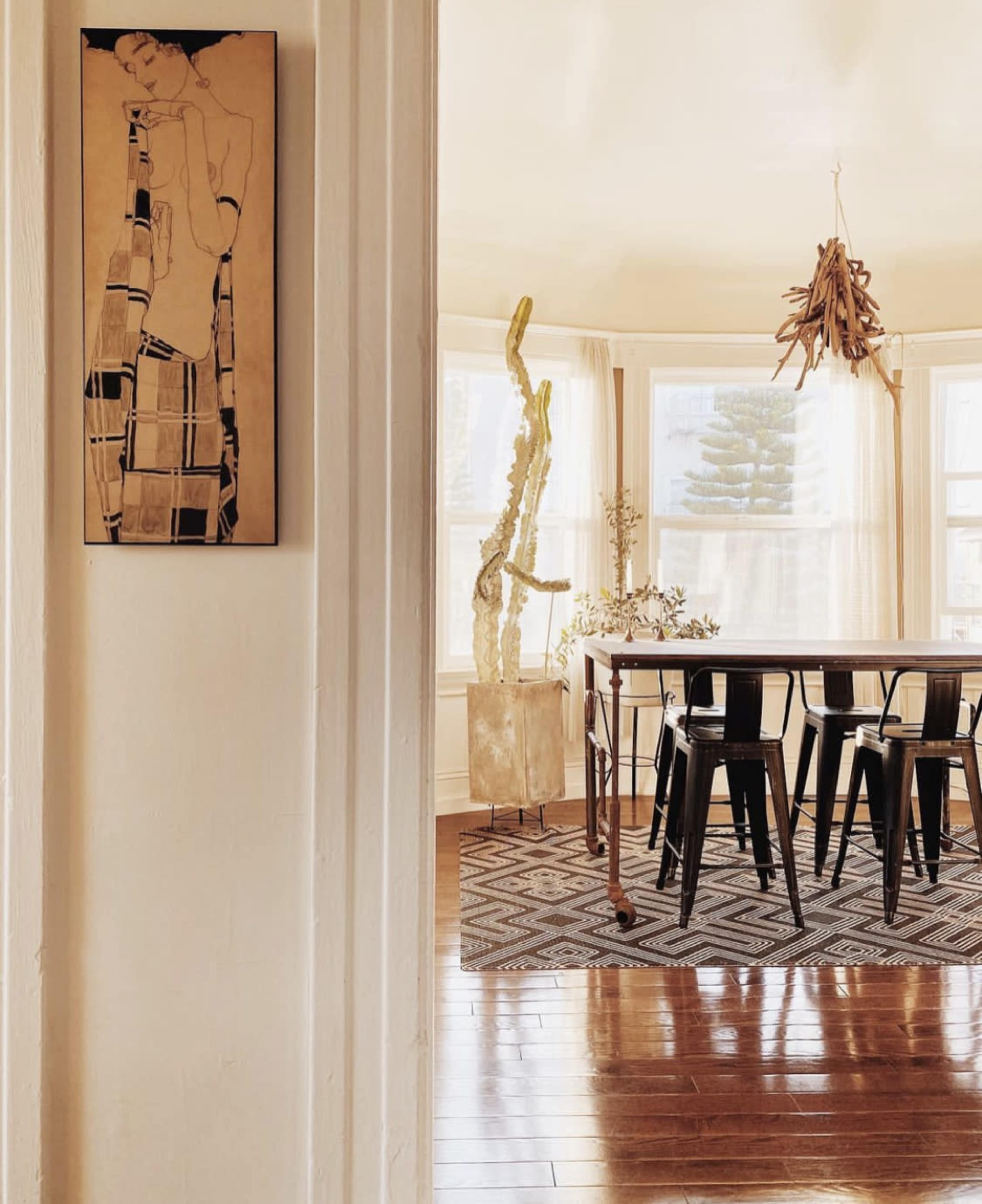 A dining area is visible through a doorway, featuring a large table surrounded by black chairs, a textured rug, and a tall plant in the corner.