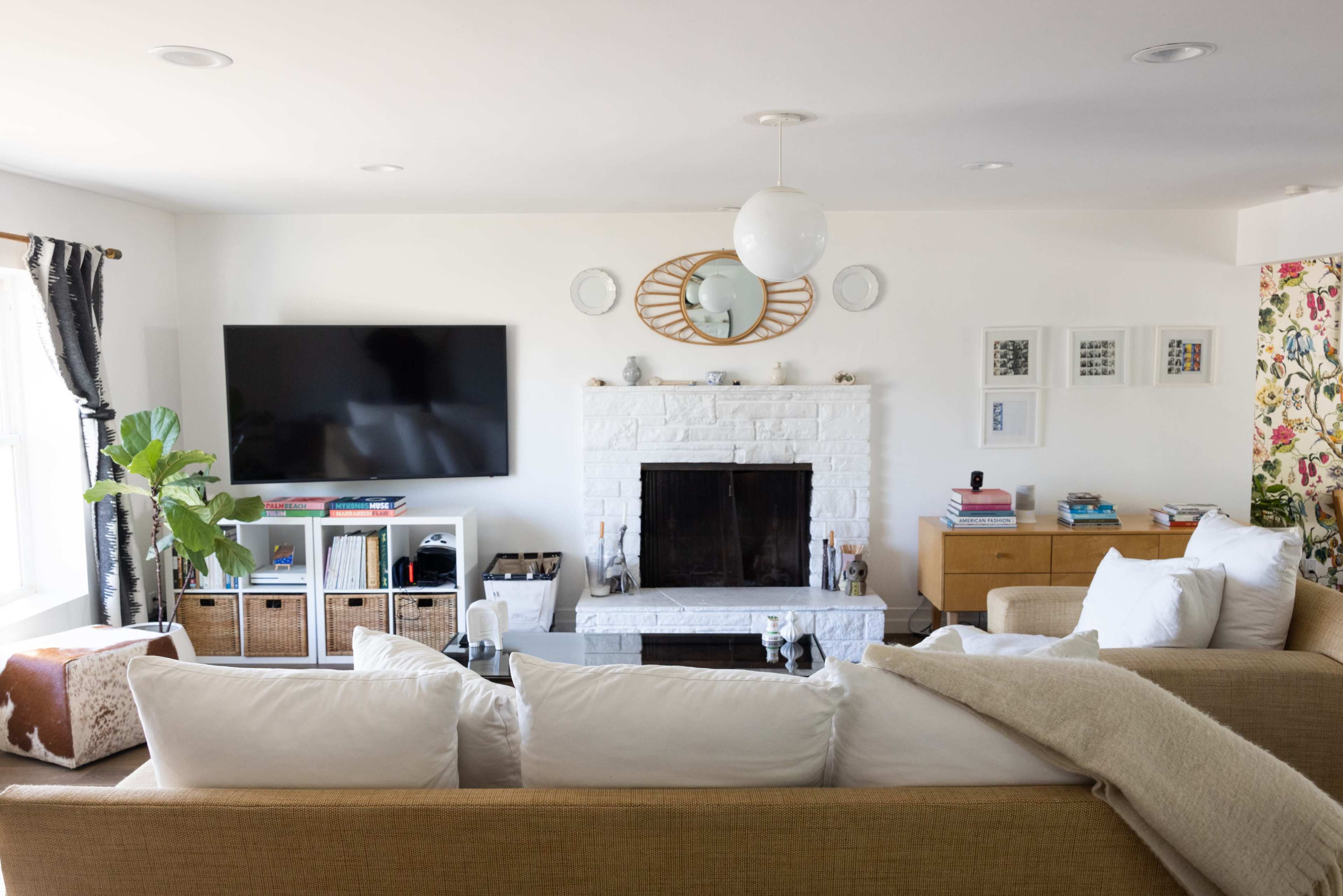 A living room features a white brick fireplace, a mounted TV, a large sectional sofa, and decorative elements including a round mirror and a plant.