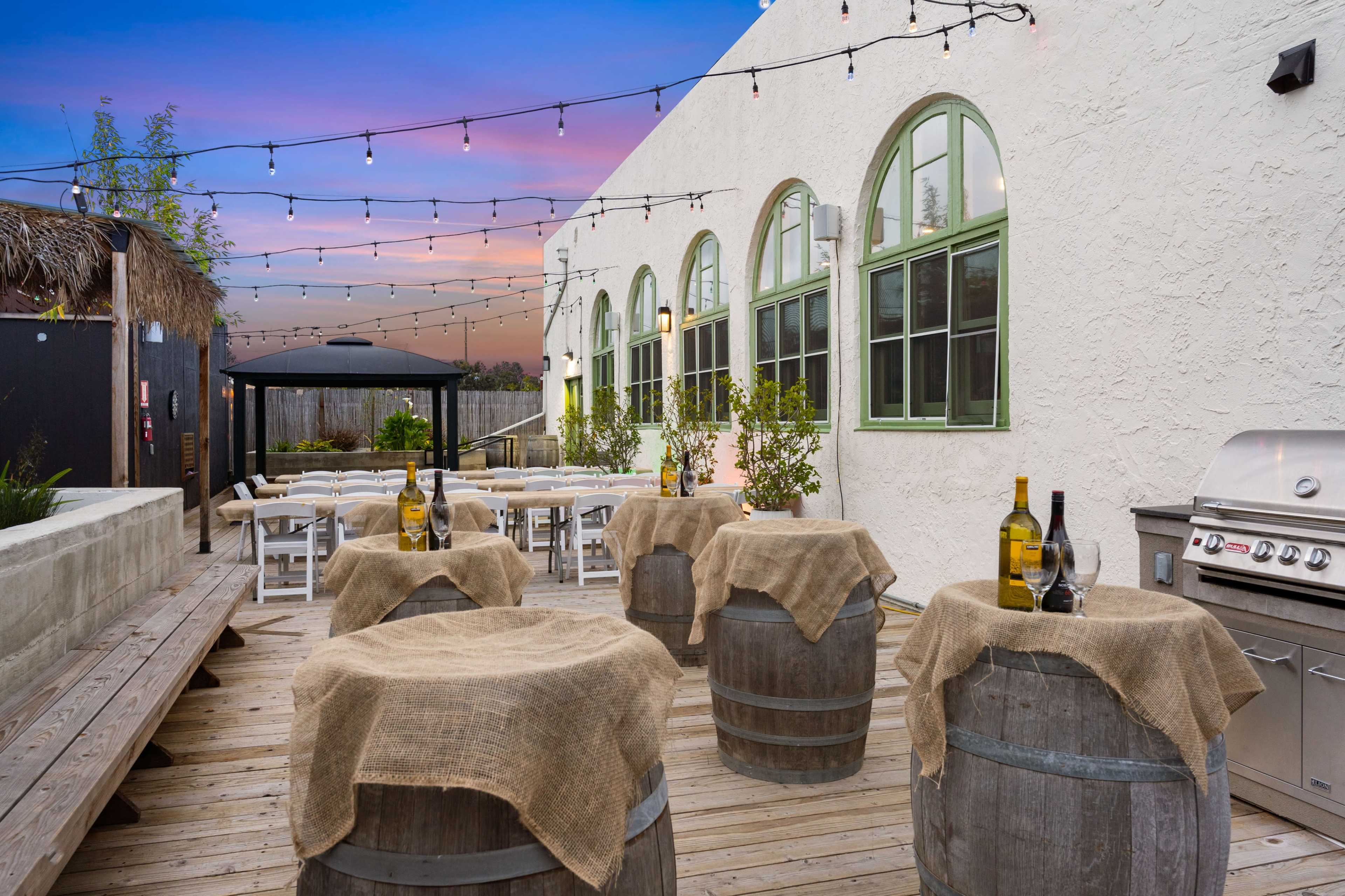 The image shows an outdoor patio area with wooden barrels topped with burlap, a barbecue grill, and tables arranged for dining under string lights against a backdrop of a colorful sunset.