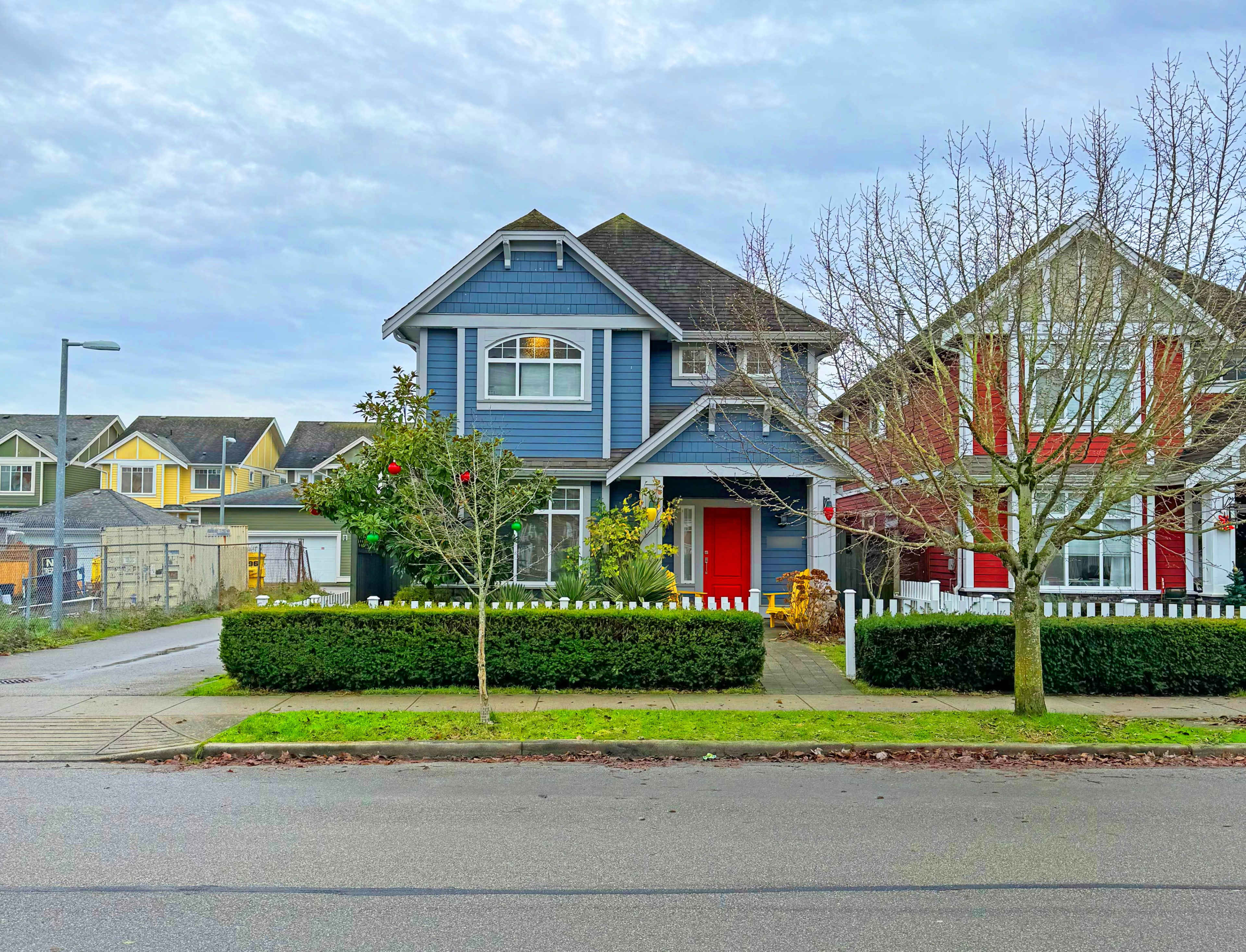 A blue two-story house with a red door is situated on a street lined with trees and a low hedge, alongside a neighboring home.