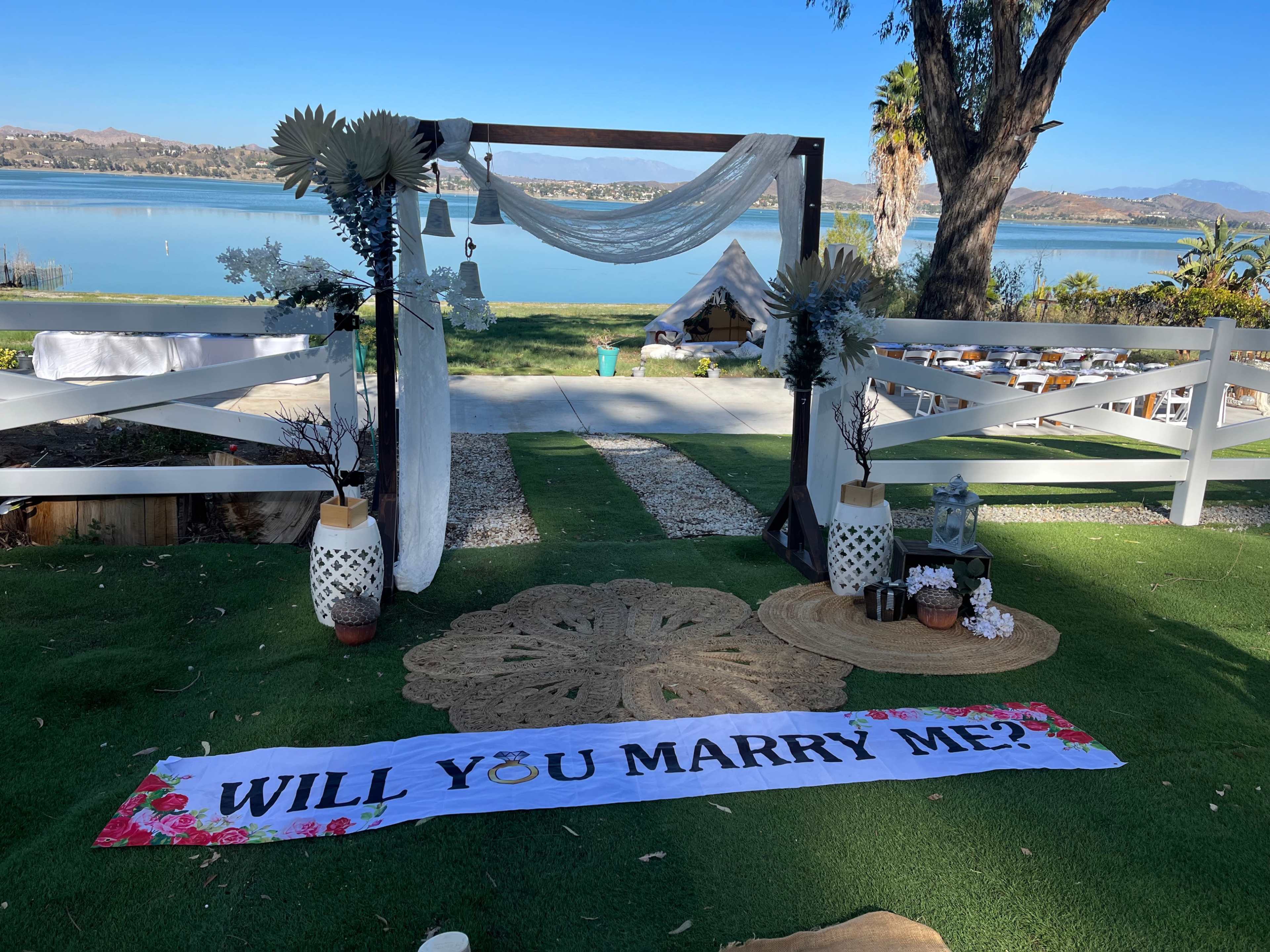 The scene features a wedding proposal setup by a lake, adorned with a decorative arch, a large floral banner reading "WILL YOU MARRY ME?", and carefully arranged decorations around a green lawn.