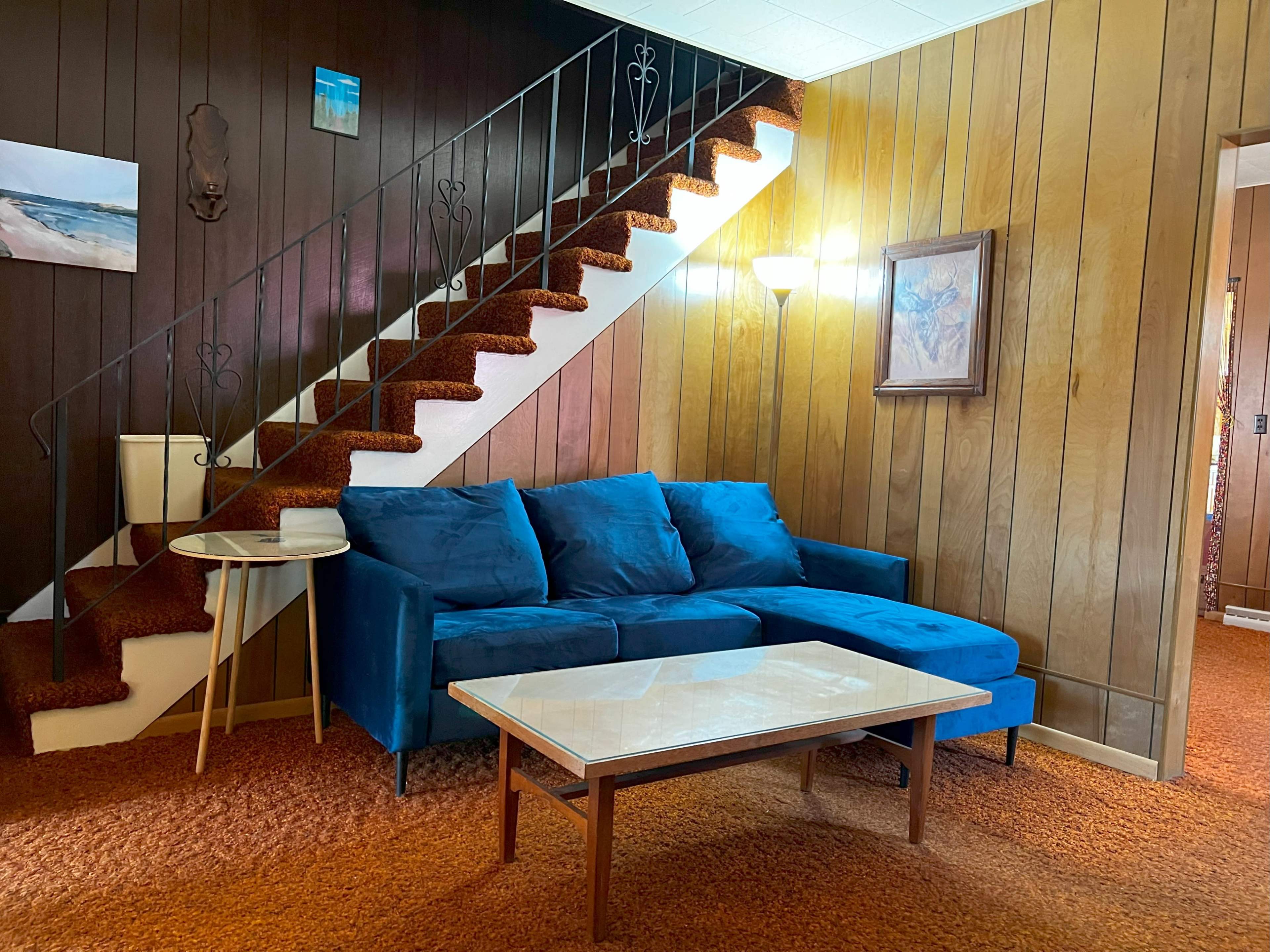 A blue sectional sofa and a marble coffee table are arranged on an orange carpeted floor beneath a staircase with decorative railings and wooden paneling on the walls.