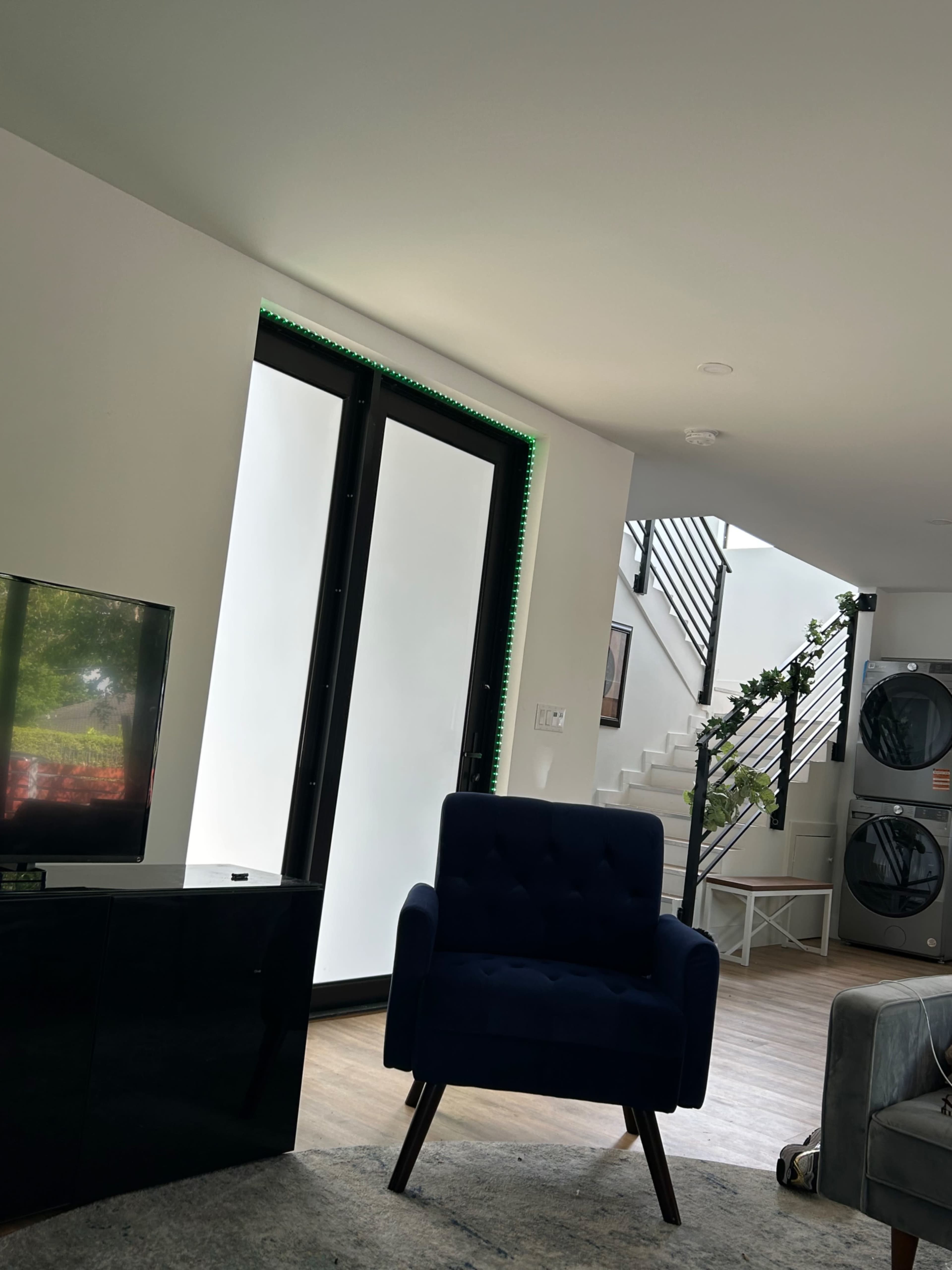 The image shows a modern living space featuring a blue armchair, a television unit, and a staircase leading to an upper level, with greenery visible near the stairs.