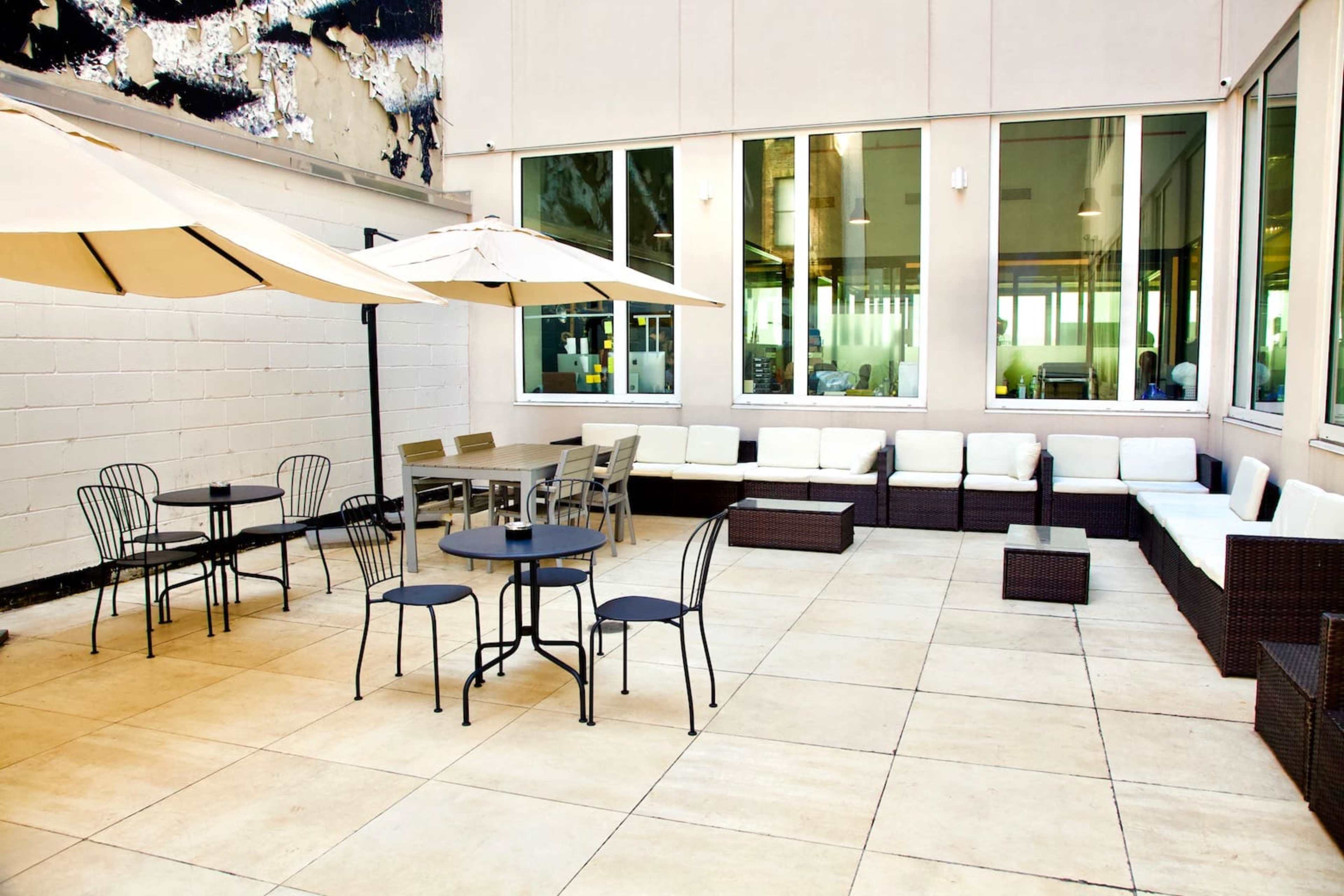 The image shows a patio area with outdoor seating, including tables and chairs under umbrellas, next to a gray wall and large windows.