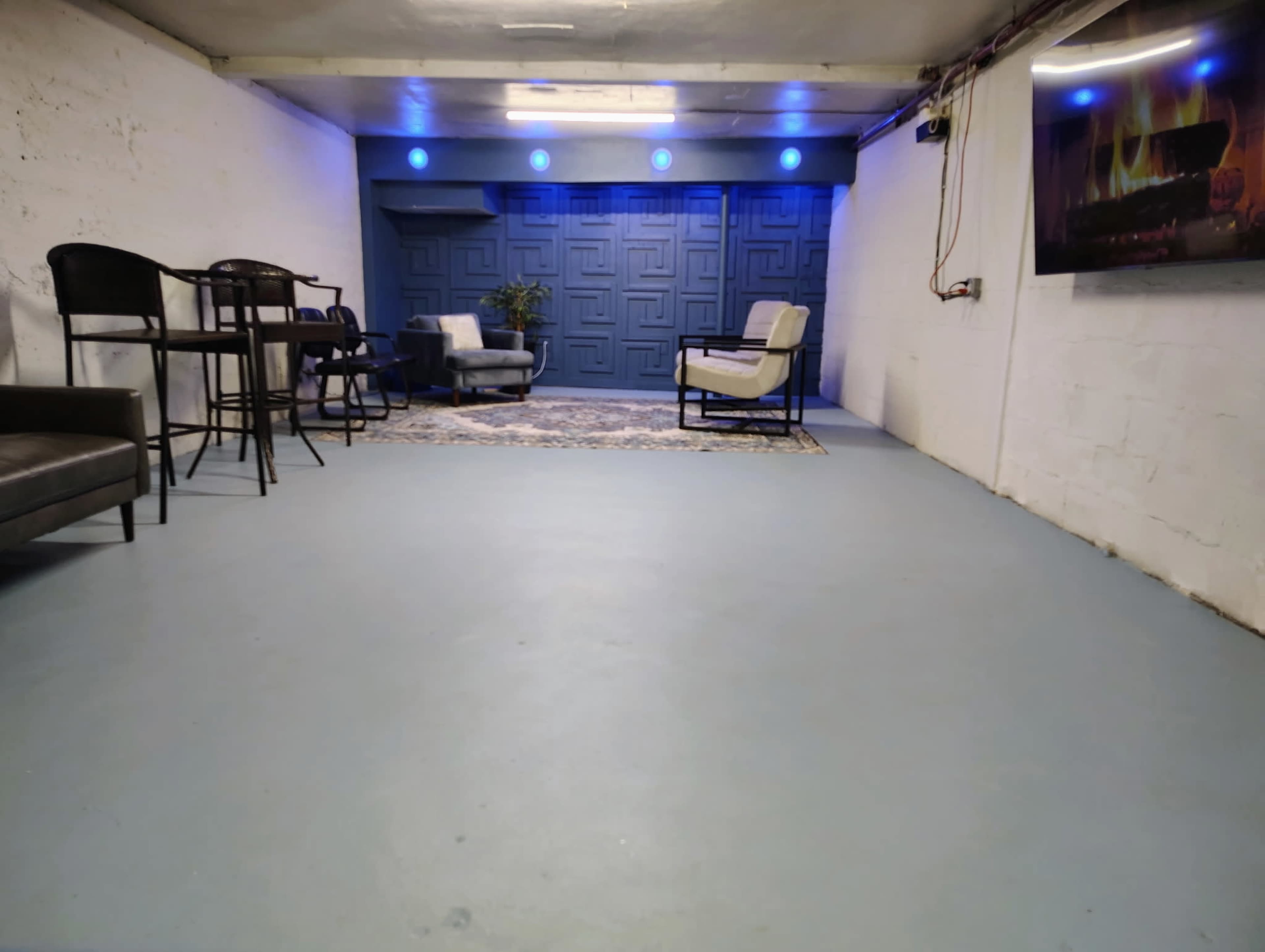 The image shows a spacious, unfinished basement featuring a seating area with two chairs, a bar table, and blue ambient lighting on the walls.