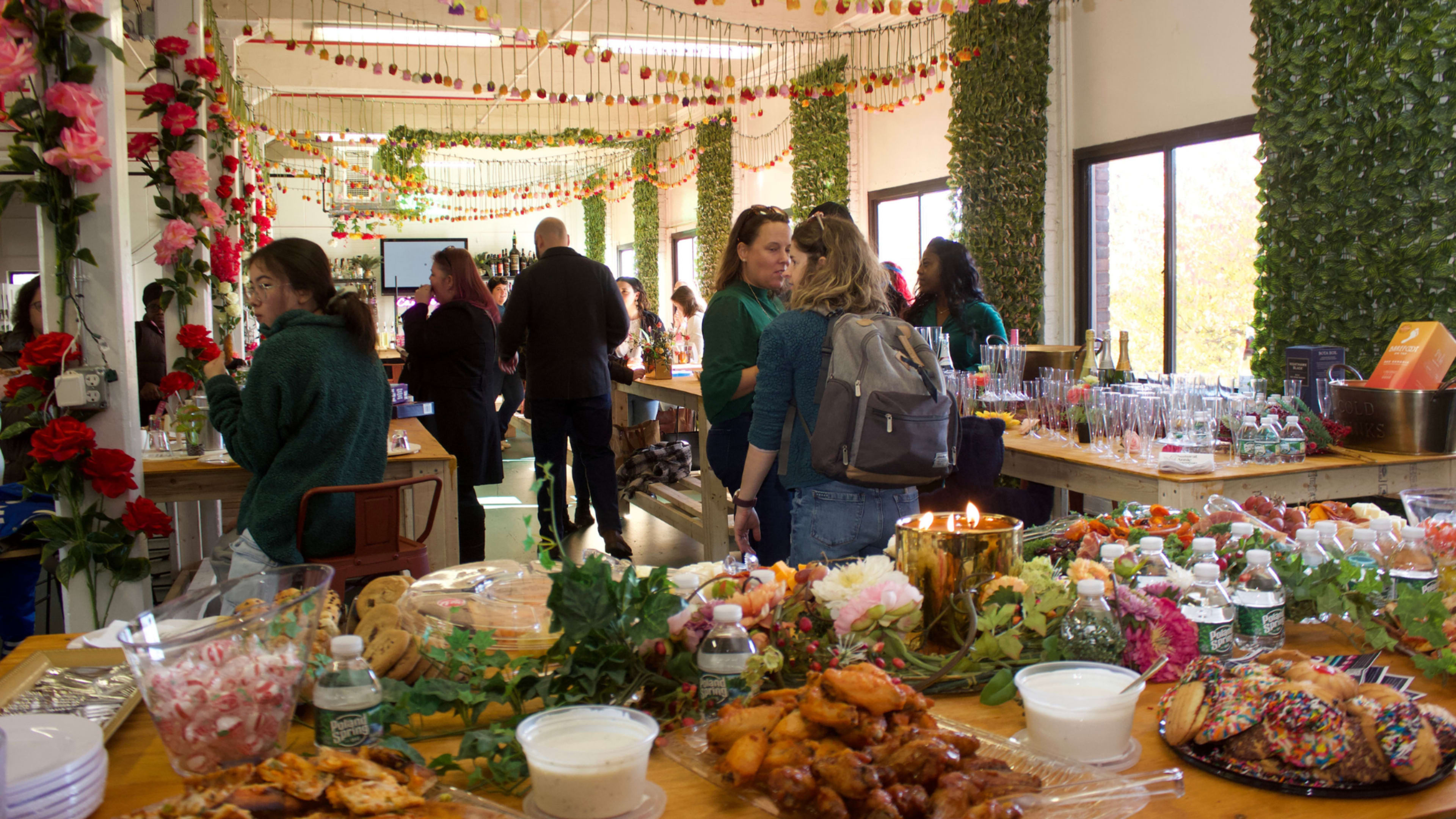 A festive gathering features people mingling around tables filled with a variety of food, surrounded by floral decorations and greenery.