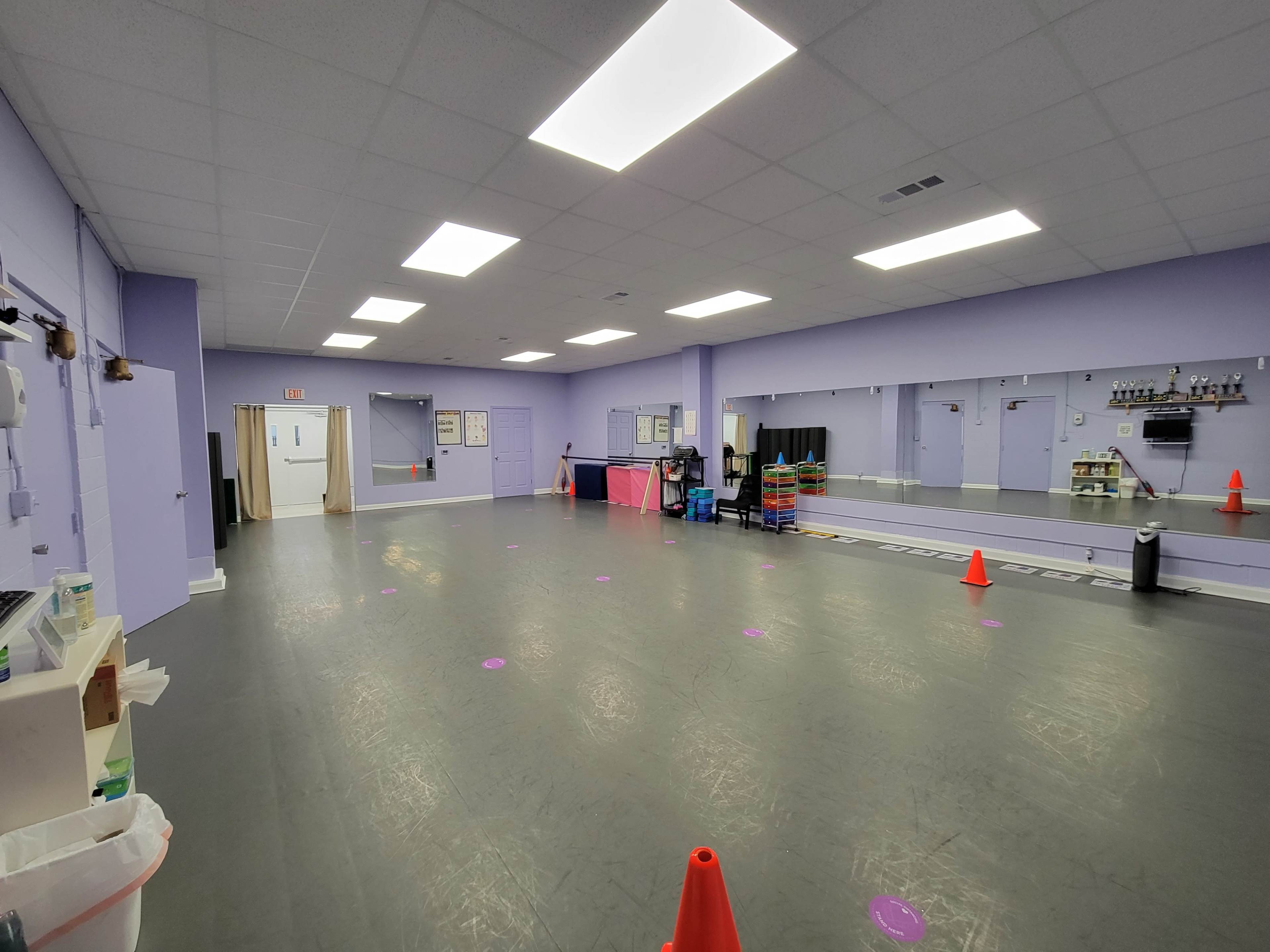 A spacious purple-walled room with a mirrored wall, featuring scattered orange cones and various equipment along one side.