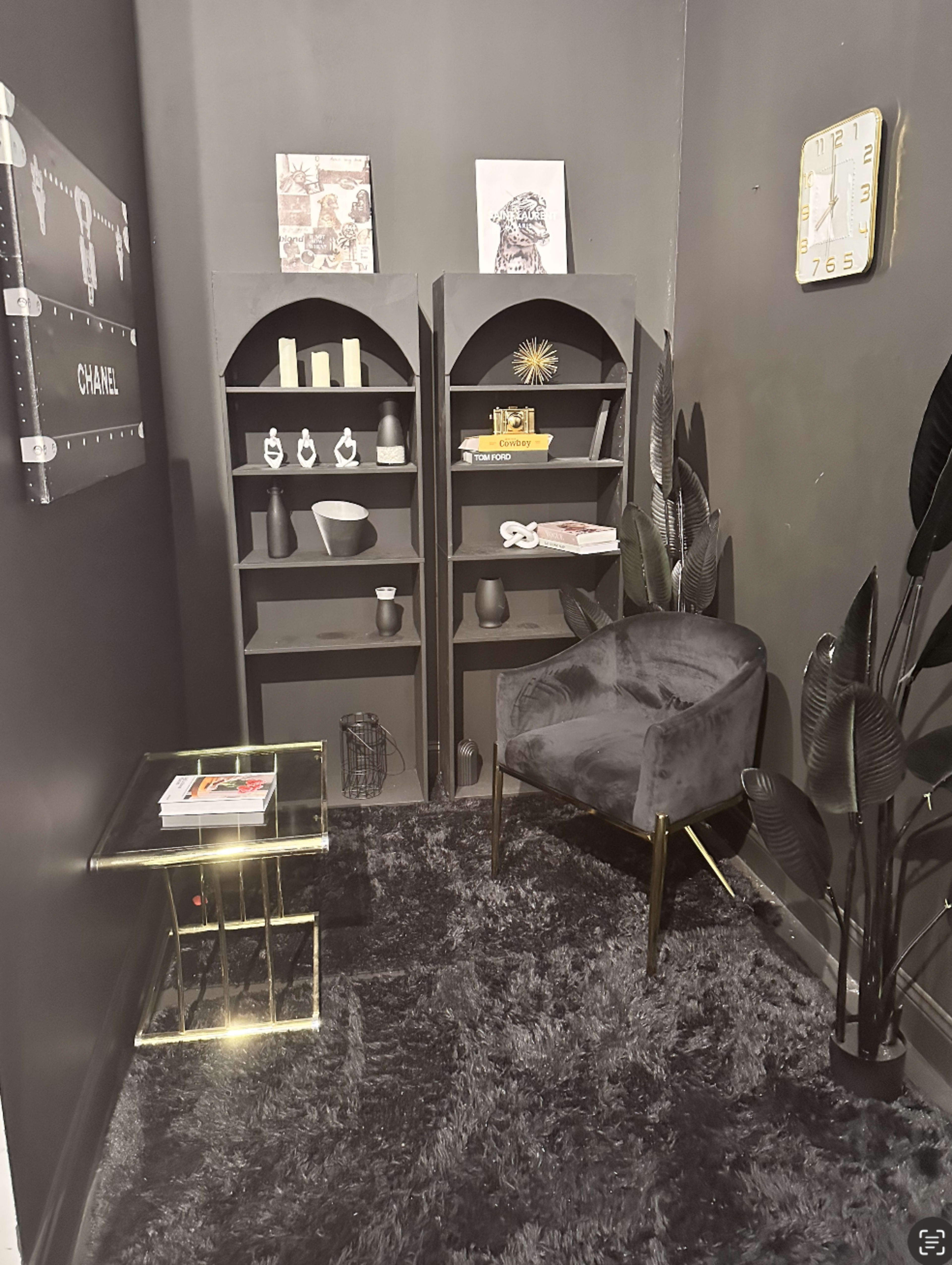 The image shows a small, dark room with a black wall, featuring a corner bookshelf, a velvet chair, a glass side table, and decorative items.
