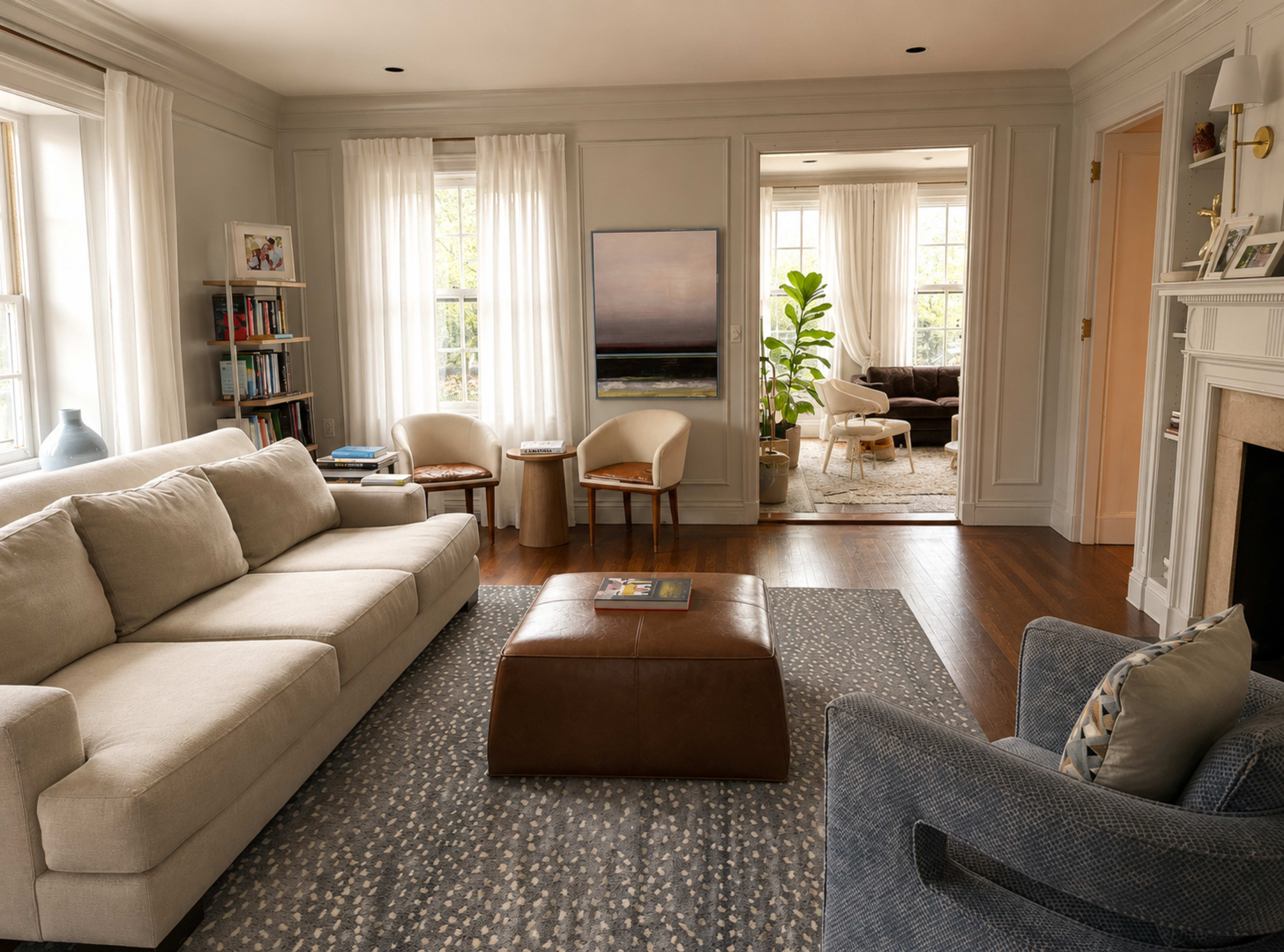 A bright living room features a light-colored sectional sofa, a brown ottoman, and adjacent seating areas with large windows and a view into another room.