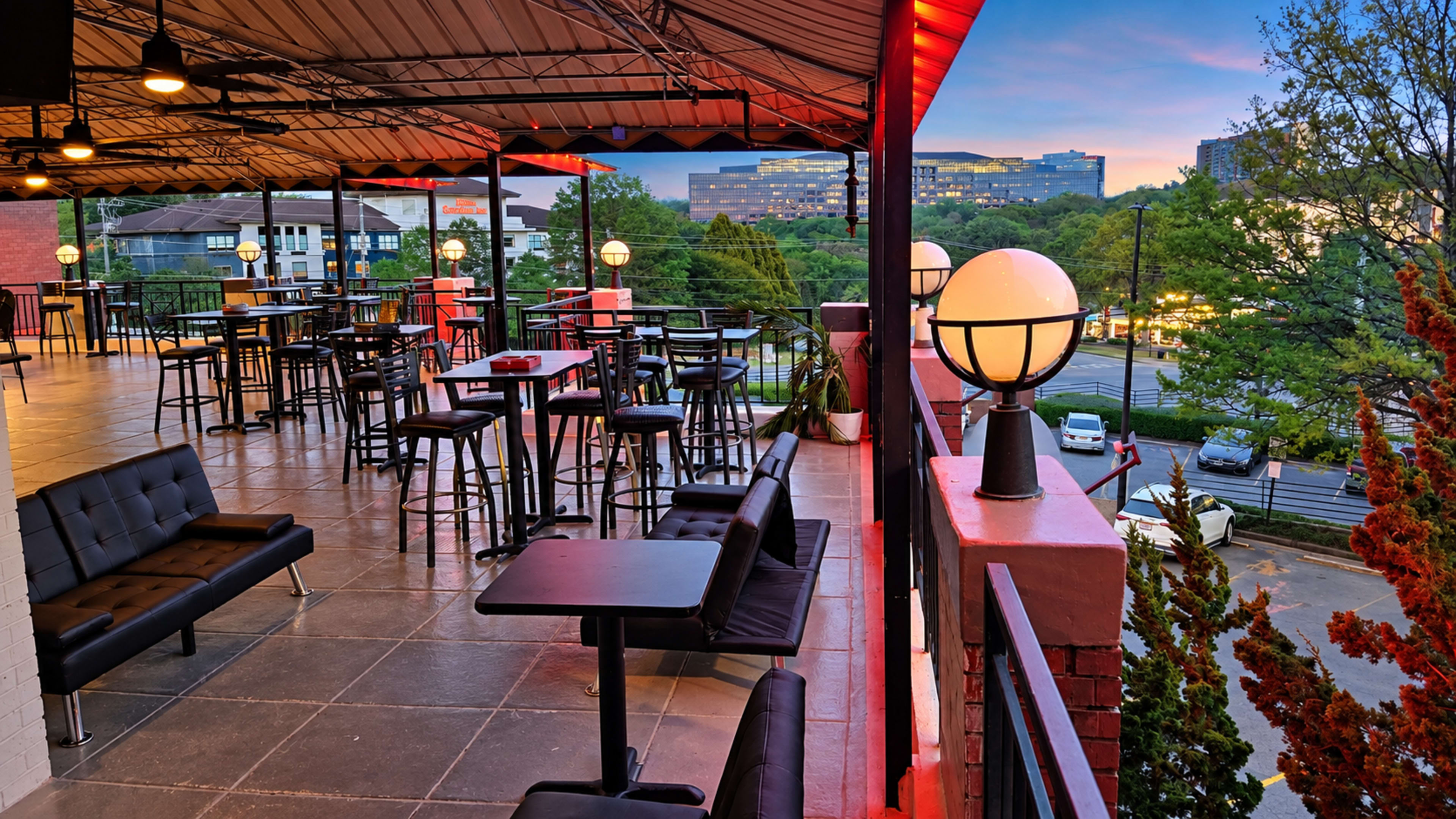 The image shows a rooftop patio with black tables and chairs, surrounded by a railing and illuminated by round lamps, overlooking a cityscape at twilight.