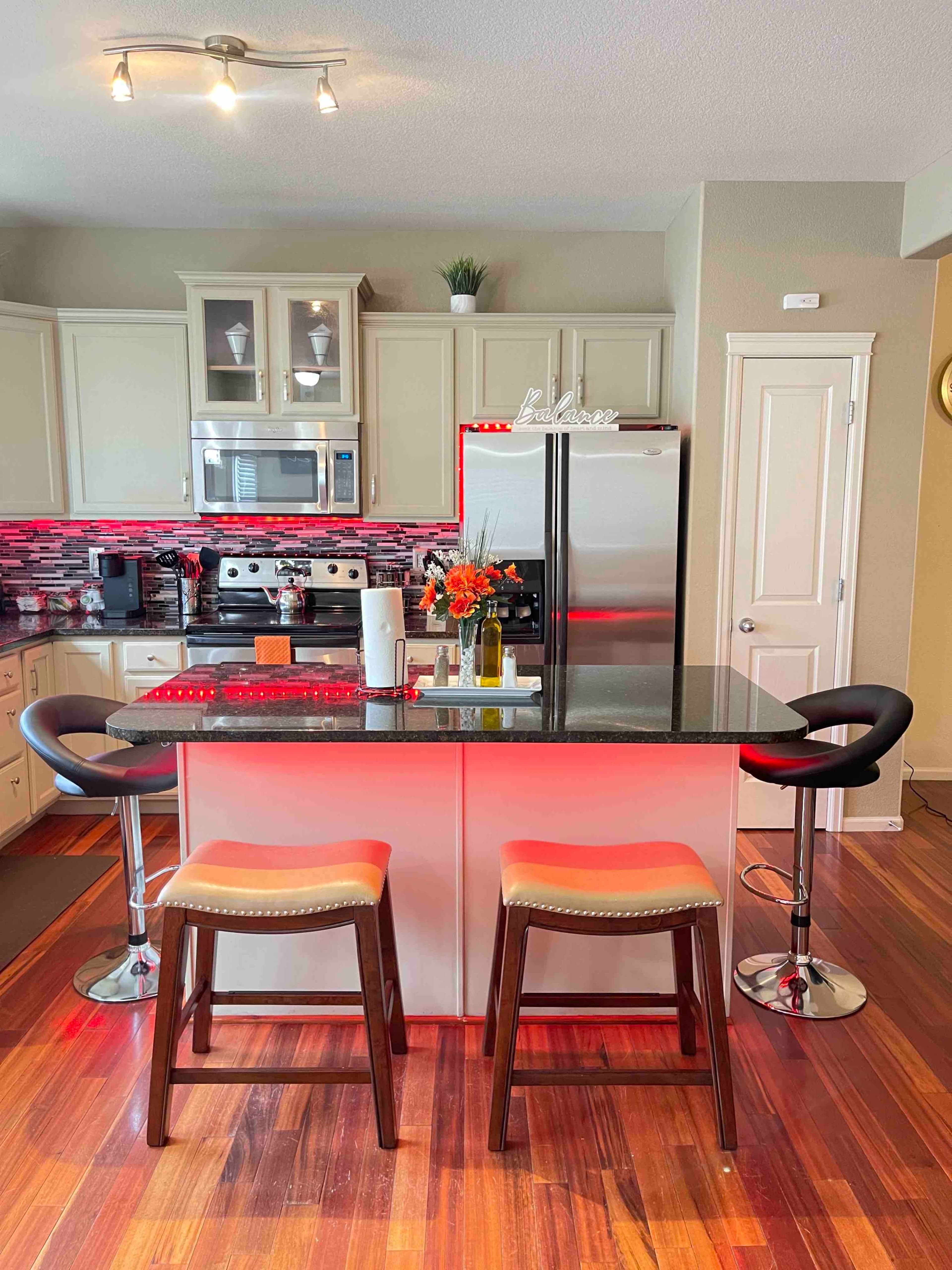 The image shows a modern kitchen with a dark countertop, stainless steel appliances, and two bar stools at a central island, illuminated by red lighting.