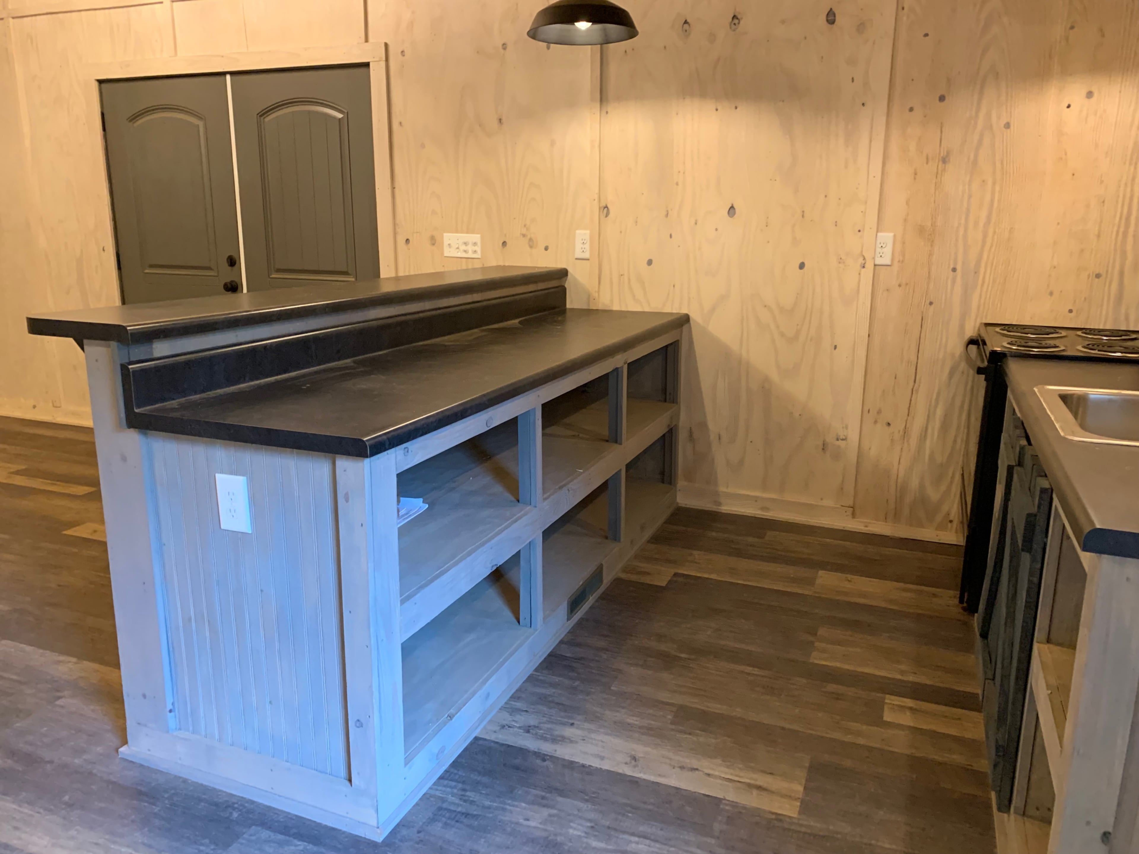 The image shows a simple kitchen with a large countertop island, open shelving, and a doorway in the background leading to another room.