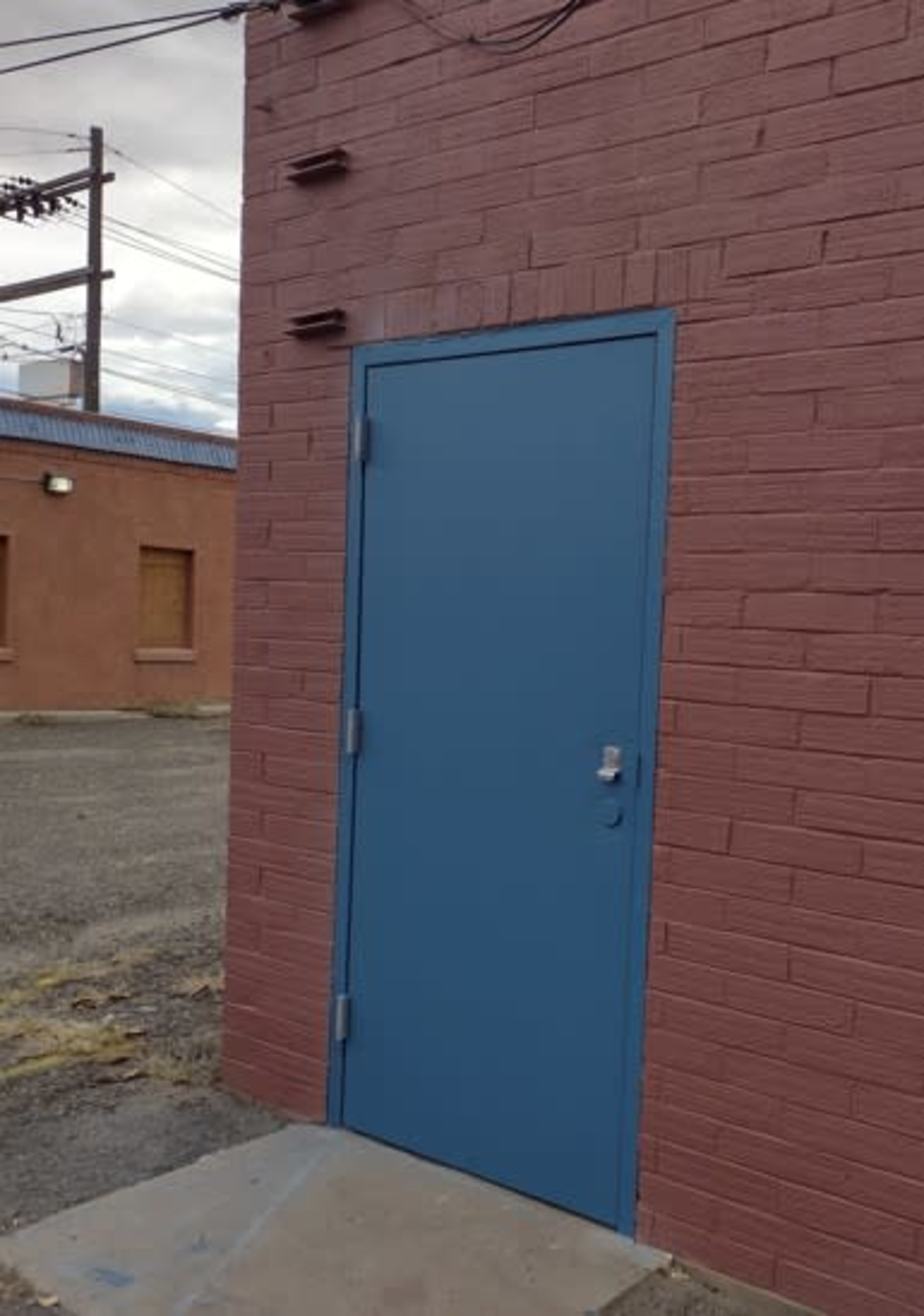 A blue door is mounted on a reddish-brown brick wall in an alleyway, with power lines visible in the background.