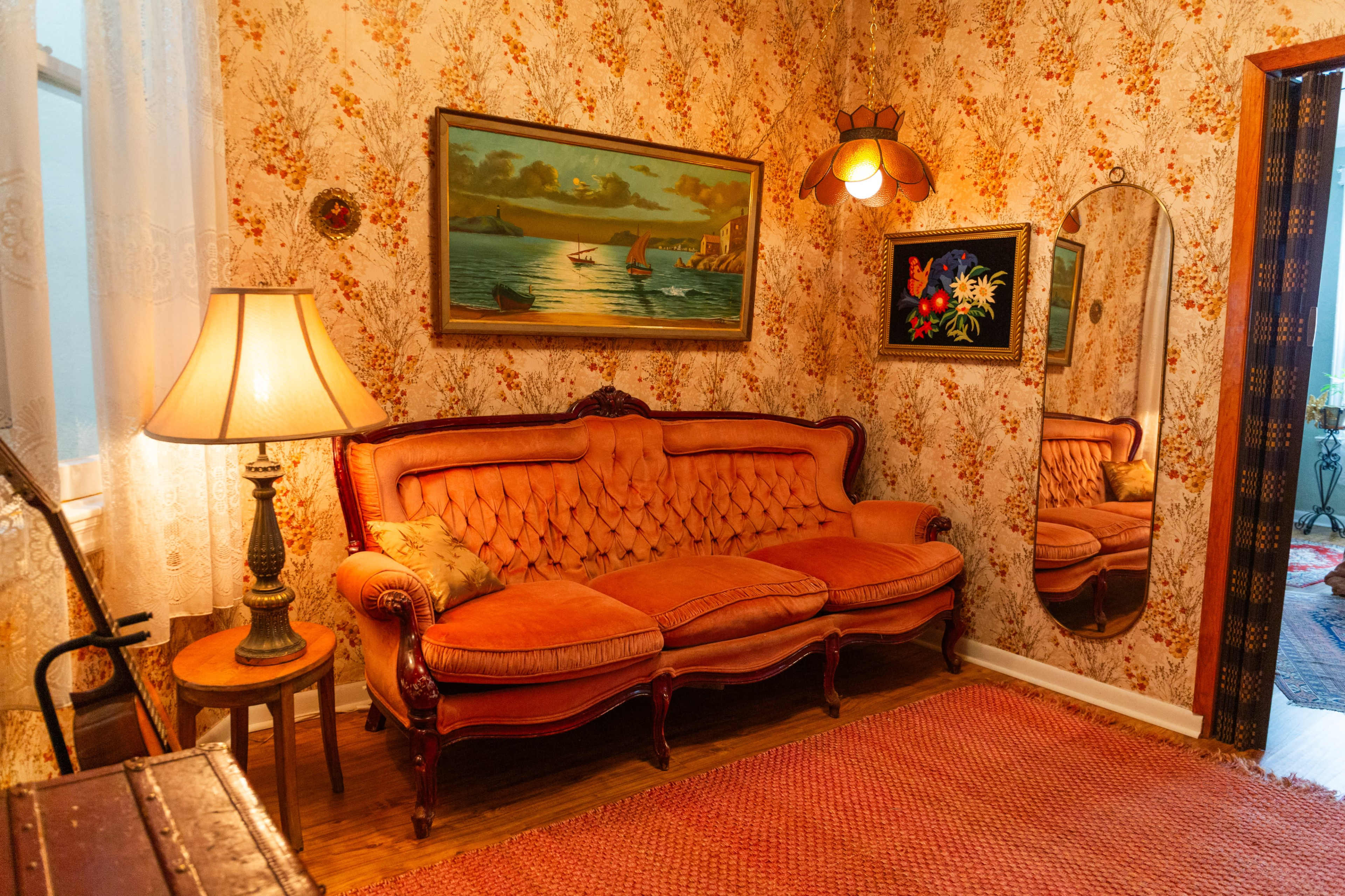 The image depicts a vintage living room with an orange tufted couch, floral wallpaper, a lamp, and paintings on the walls.