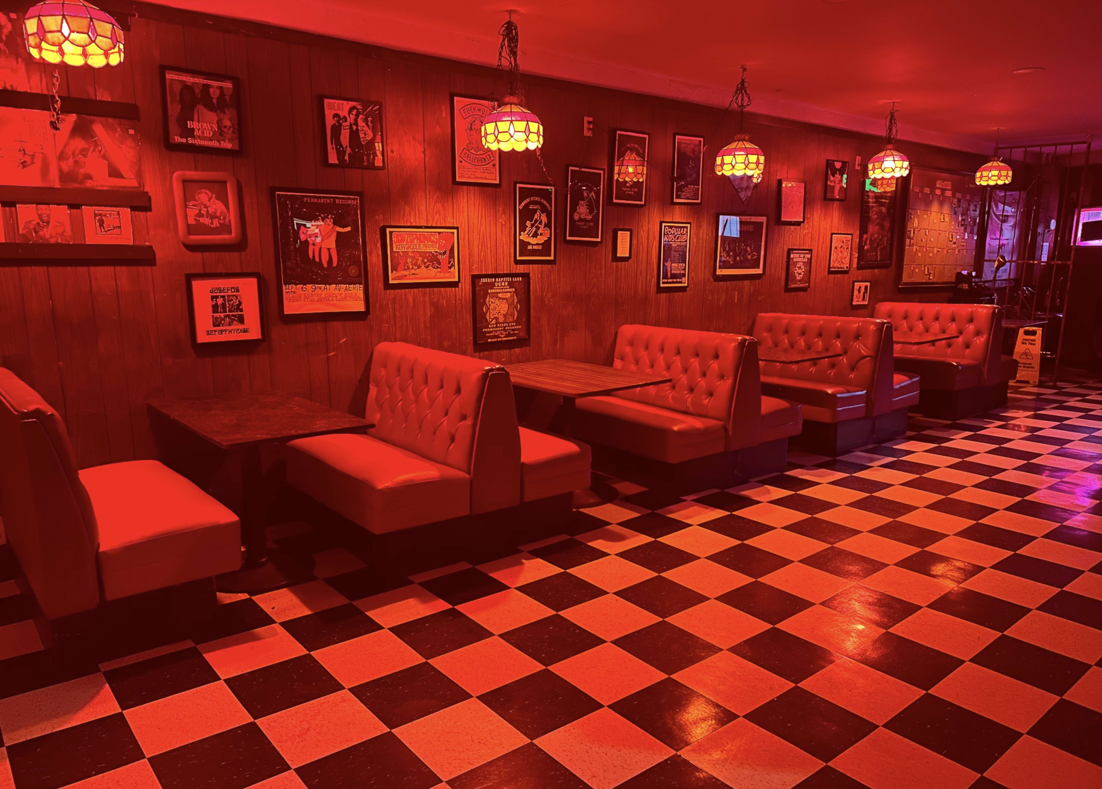 The interior of a restaurant features red upholstered booths, wooden walls adorned with framed artworks, and a black-and-white checkered floor under warm red lighting.