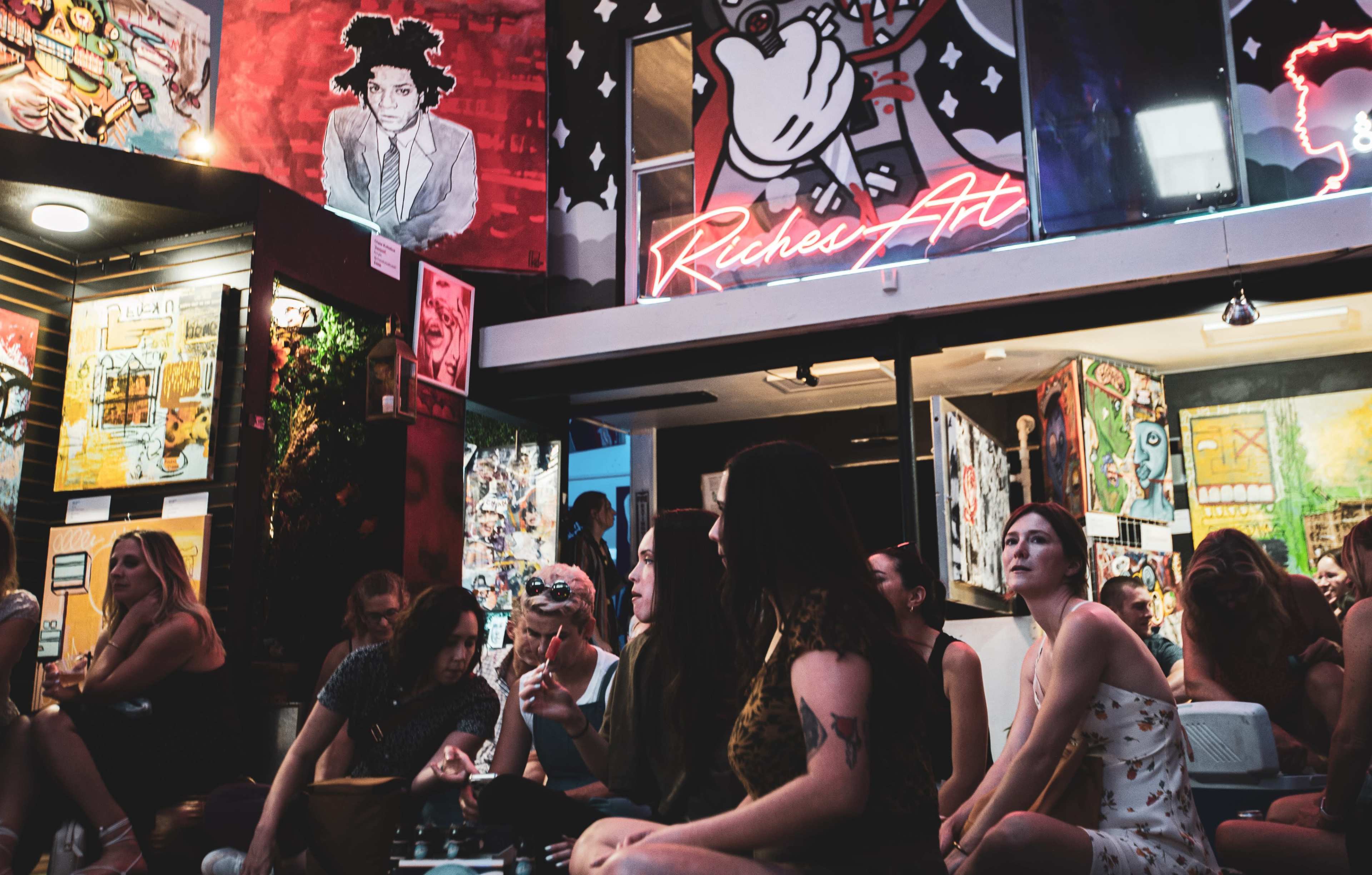 A group of people sits inside an art space surrounded by colorful murals and neon signs.