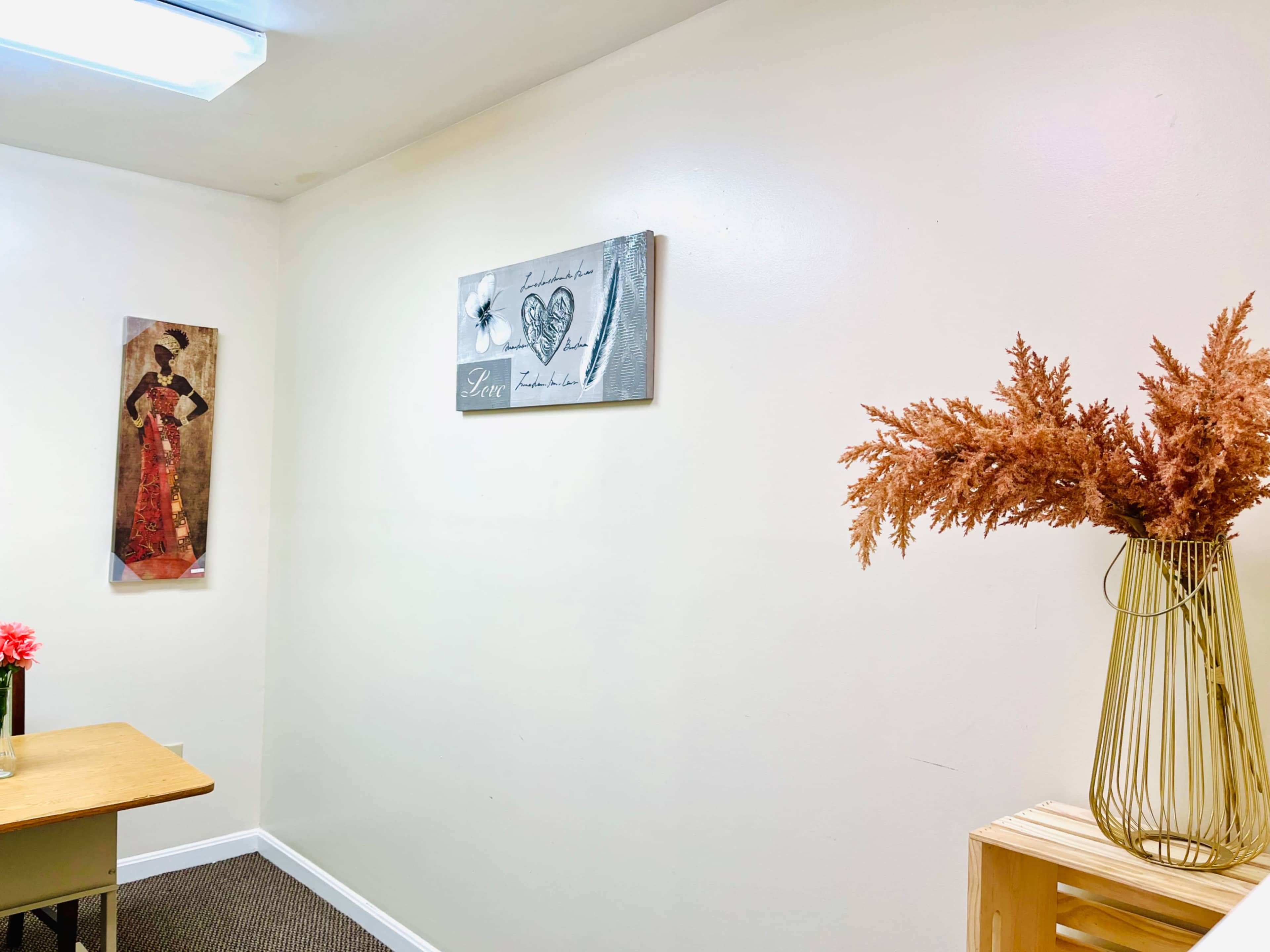A small, simple room with a table, a wall decoration featuring a figure in traditional attire, and a vase of dried orange flowers.