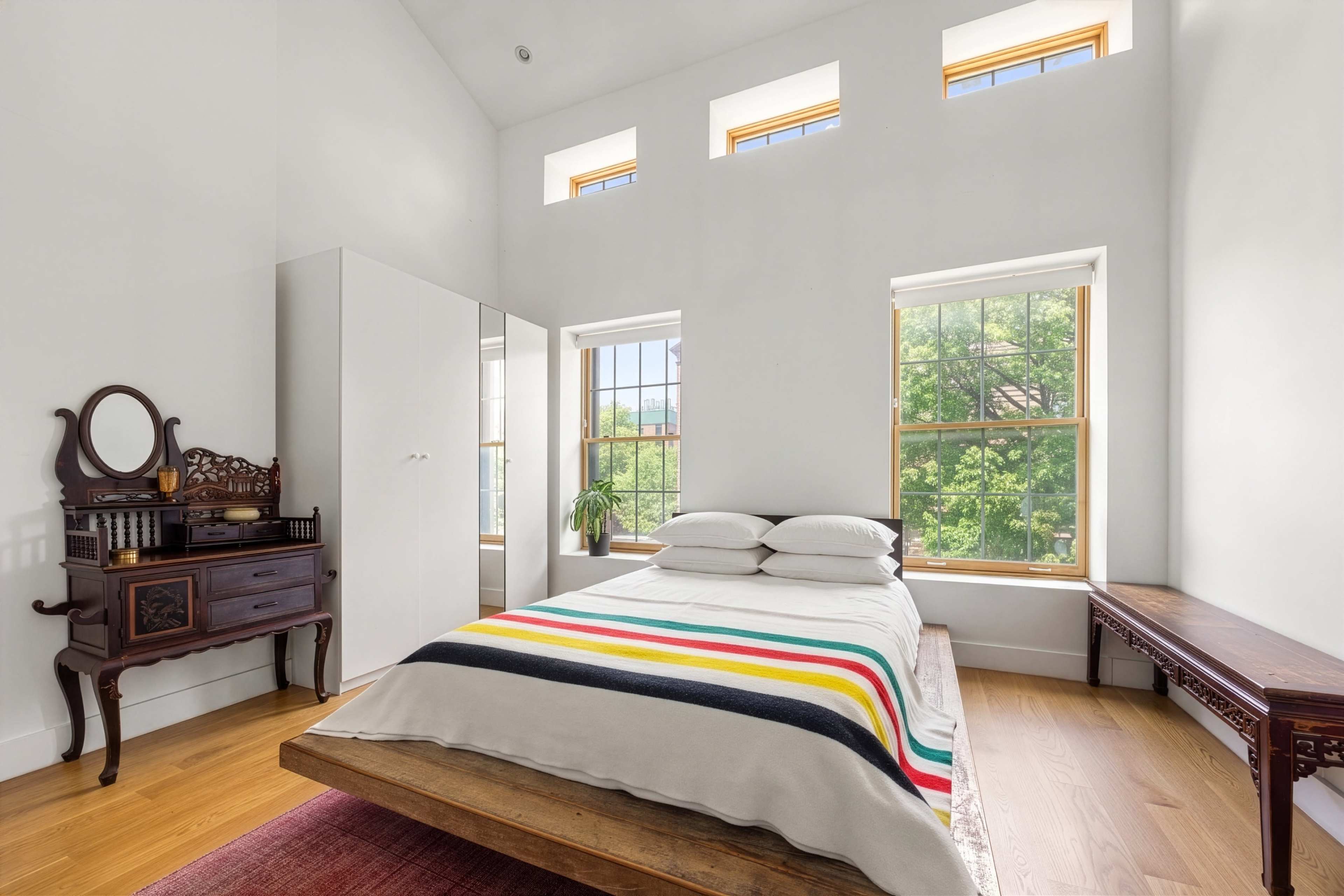 The image shows a bright bedroom with large windows, a wooden bed with a striped blanket, and a mix of modern and antique furniture.