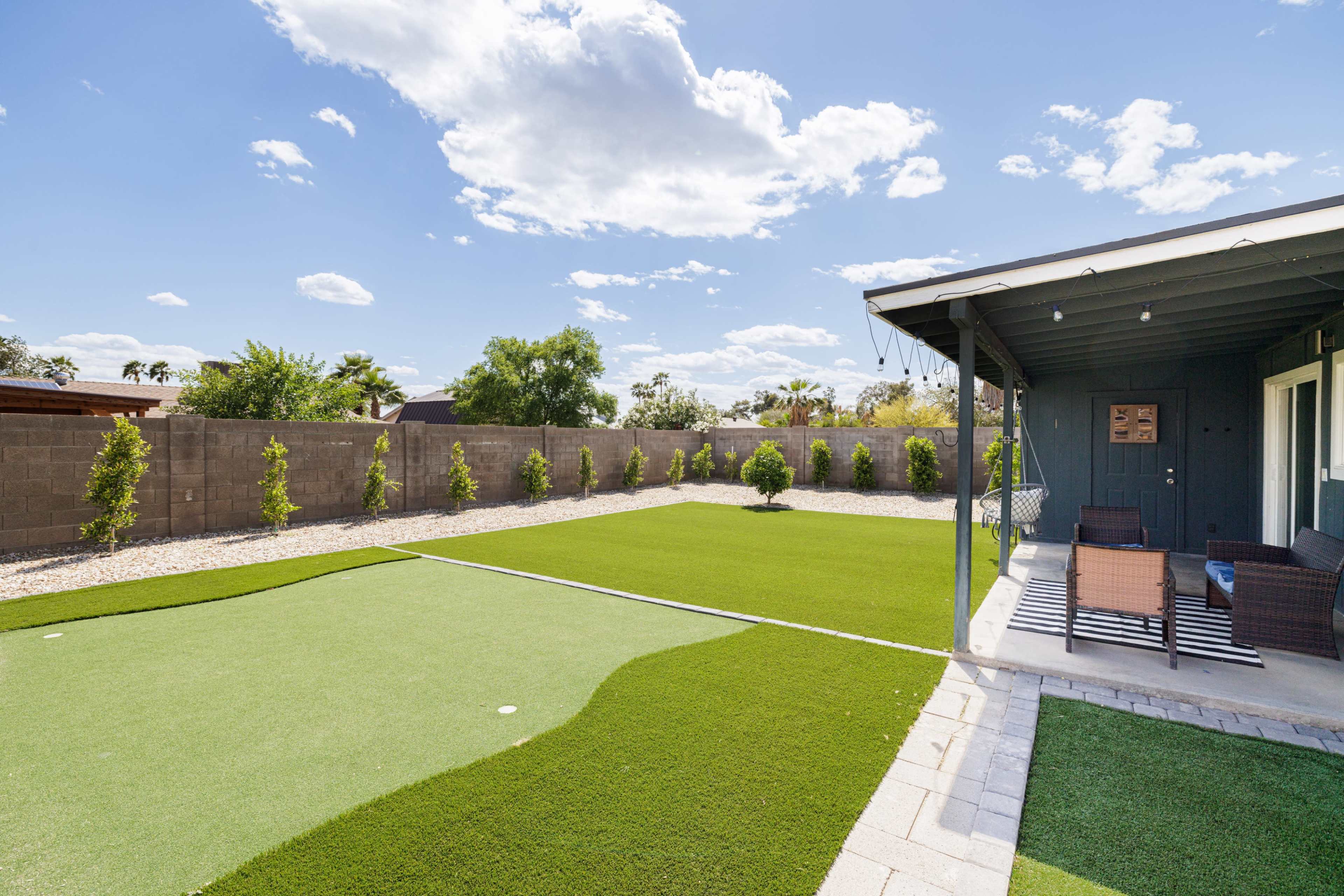 The image shows a landscaped backyard with artificial turf, bordered by low shrubs and a patio area with seating.