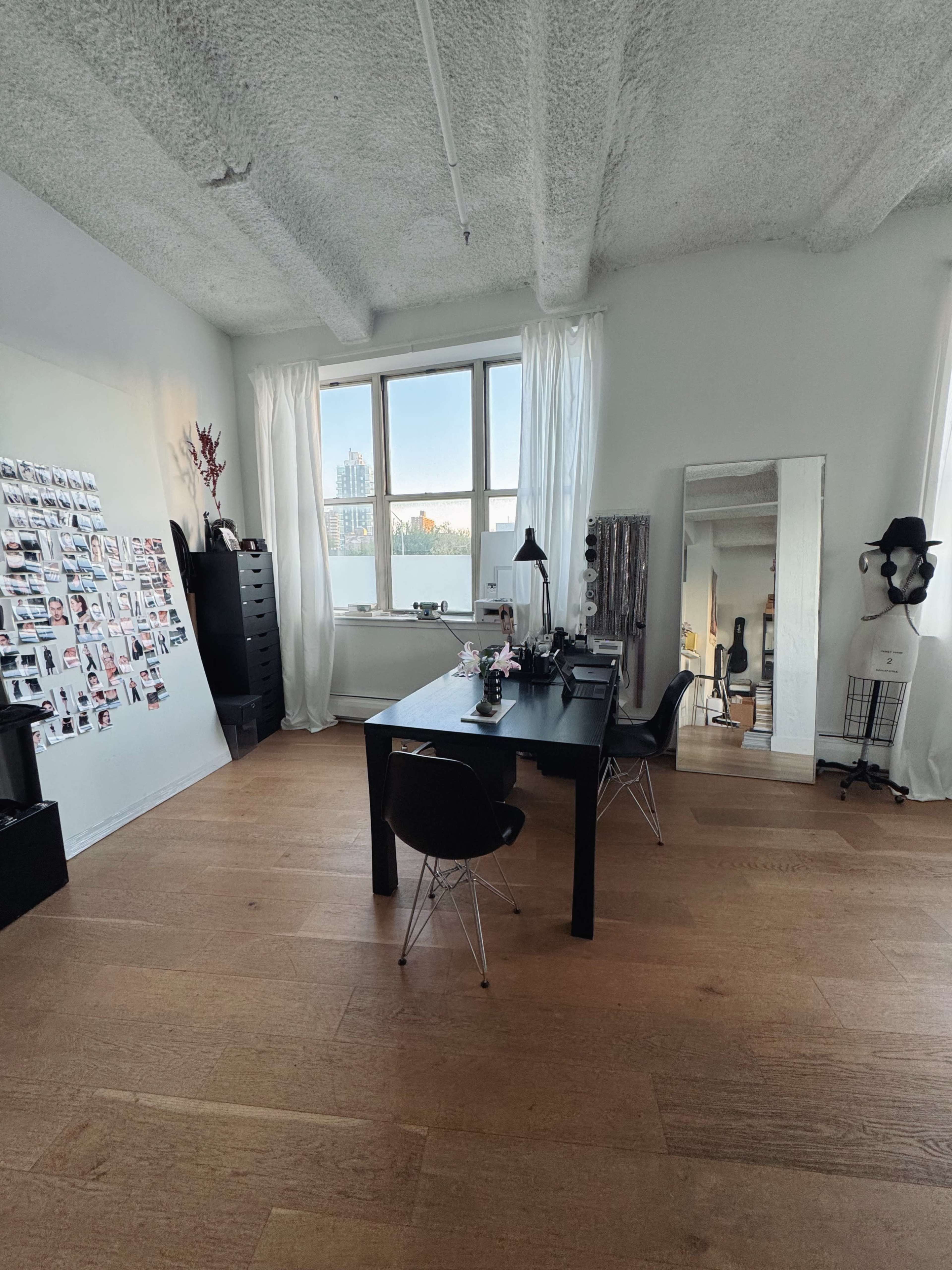 The image shows a modern office space featuring a black desk with two chairs, a large mirror, a wall decorated with photos, and natural light coming through a window.