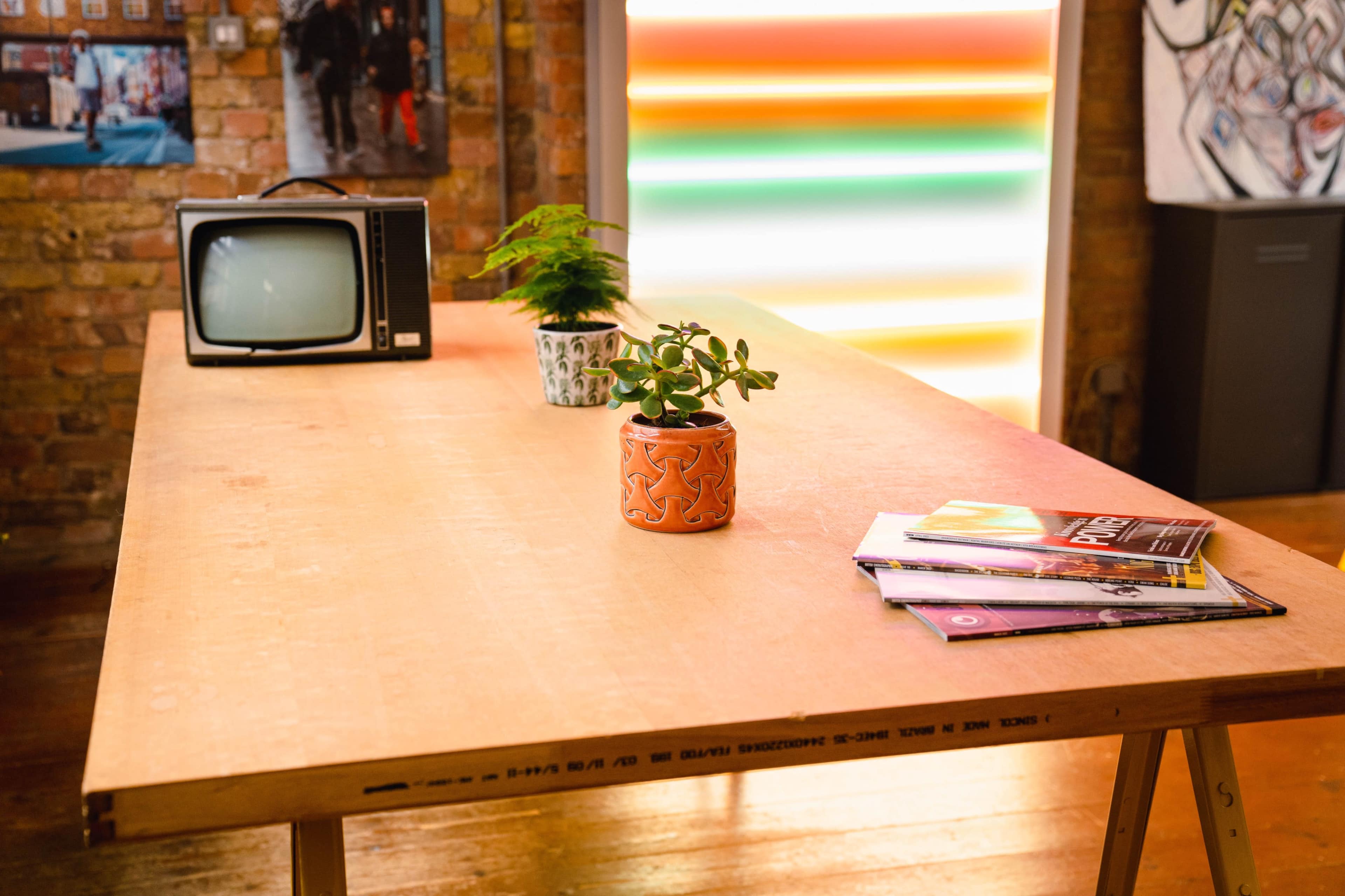 A wooden table displays a vintage television, a potted plant, and a stack of magazines in a well-lit room with brick walls.