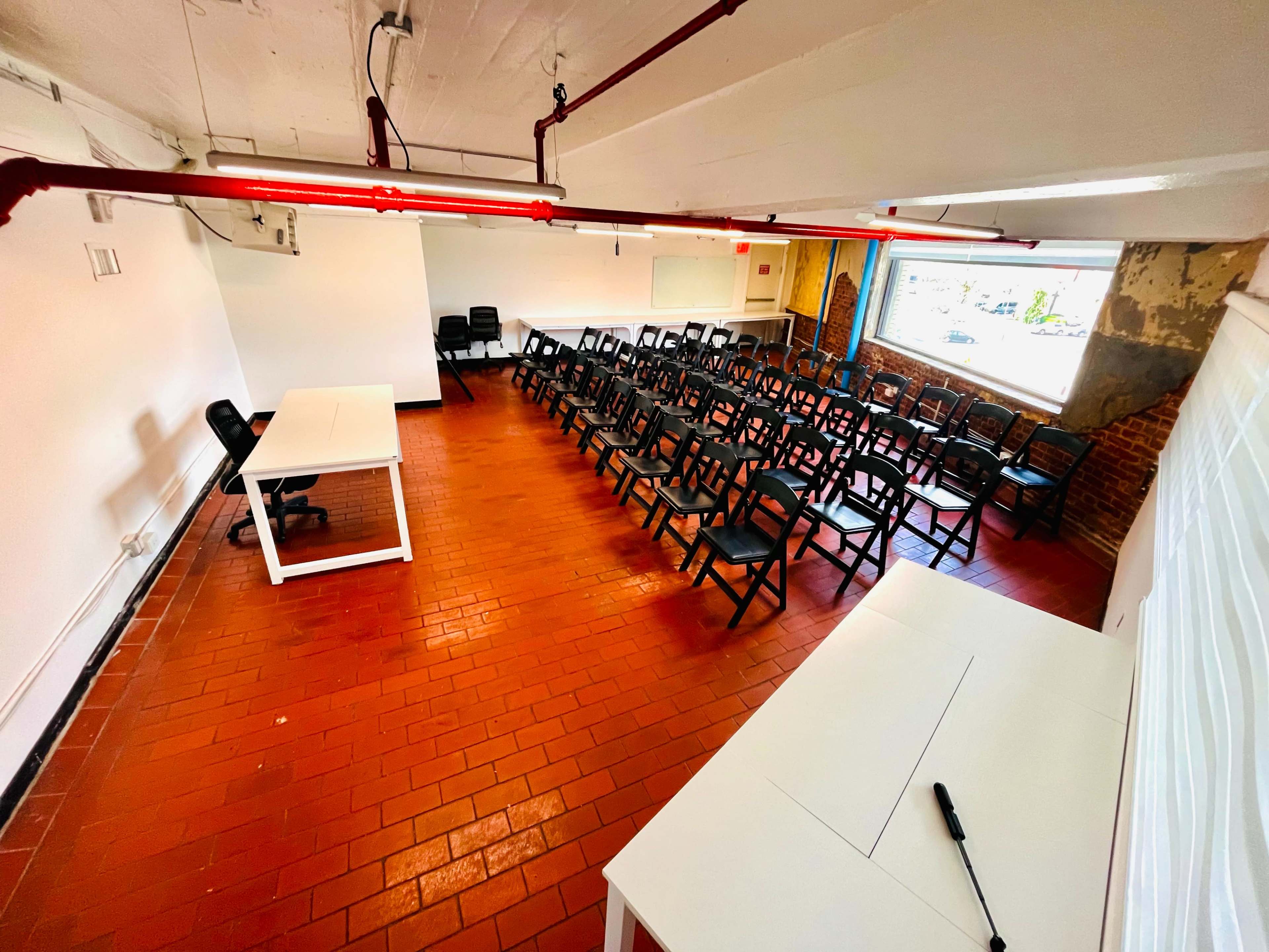A spacious classroom features rows of black chairs facing a whiteboard, with a large window letting in natural light.