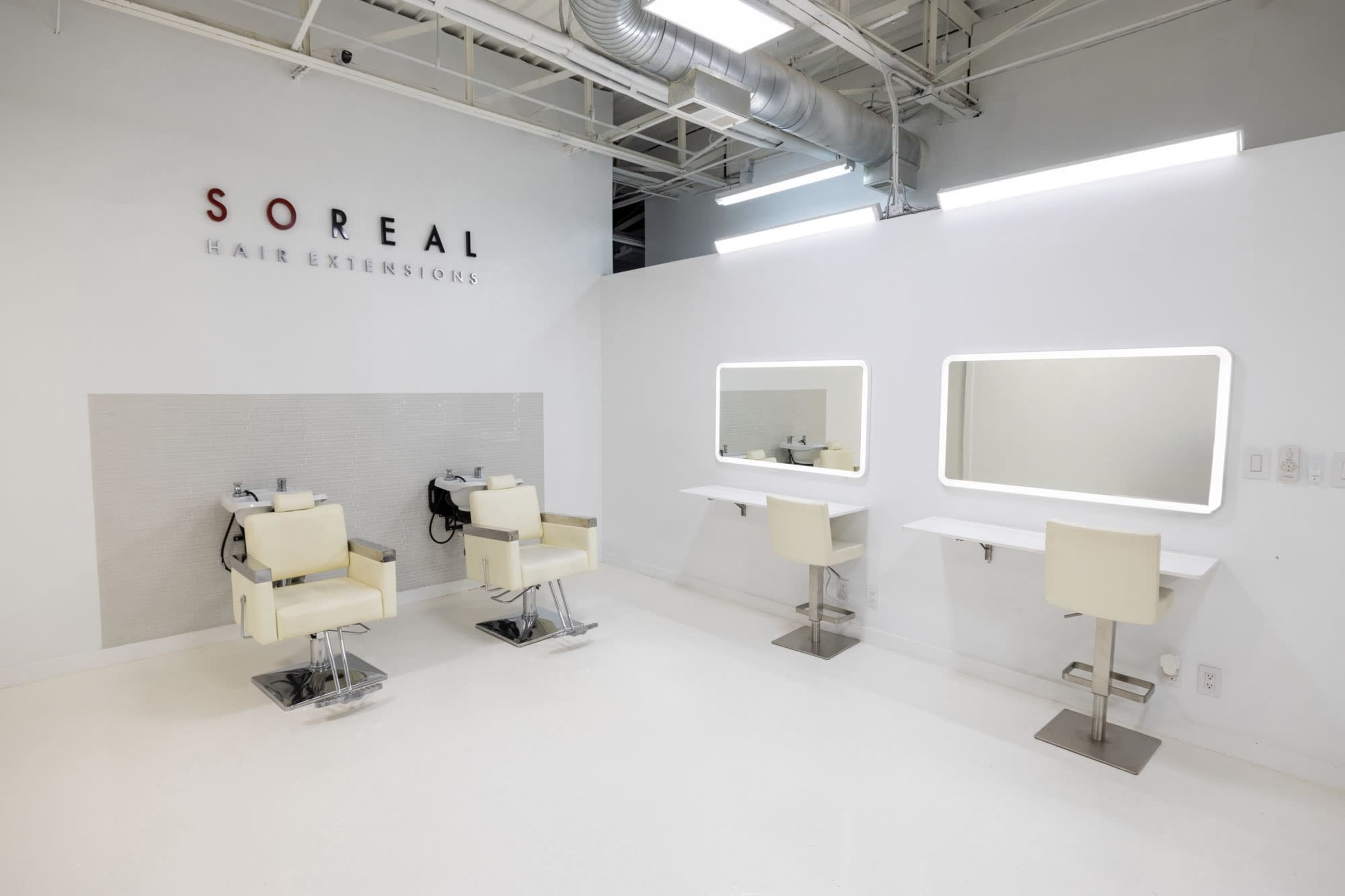 The image shows a minimalist hair salon with two styling chairs, mirrors, and a sleek, white interior.
