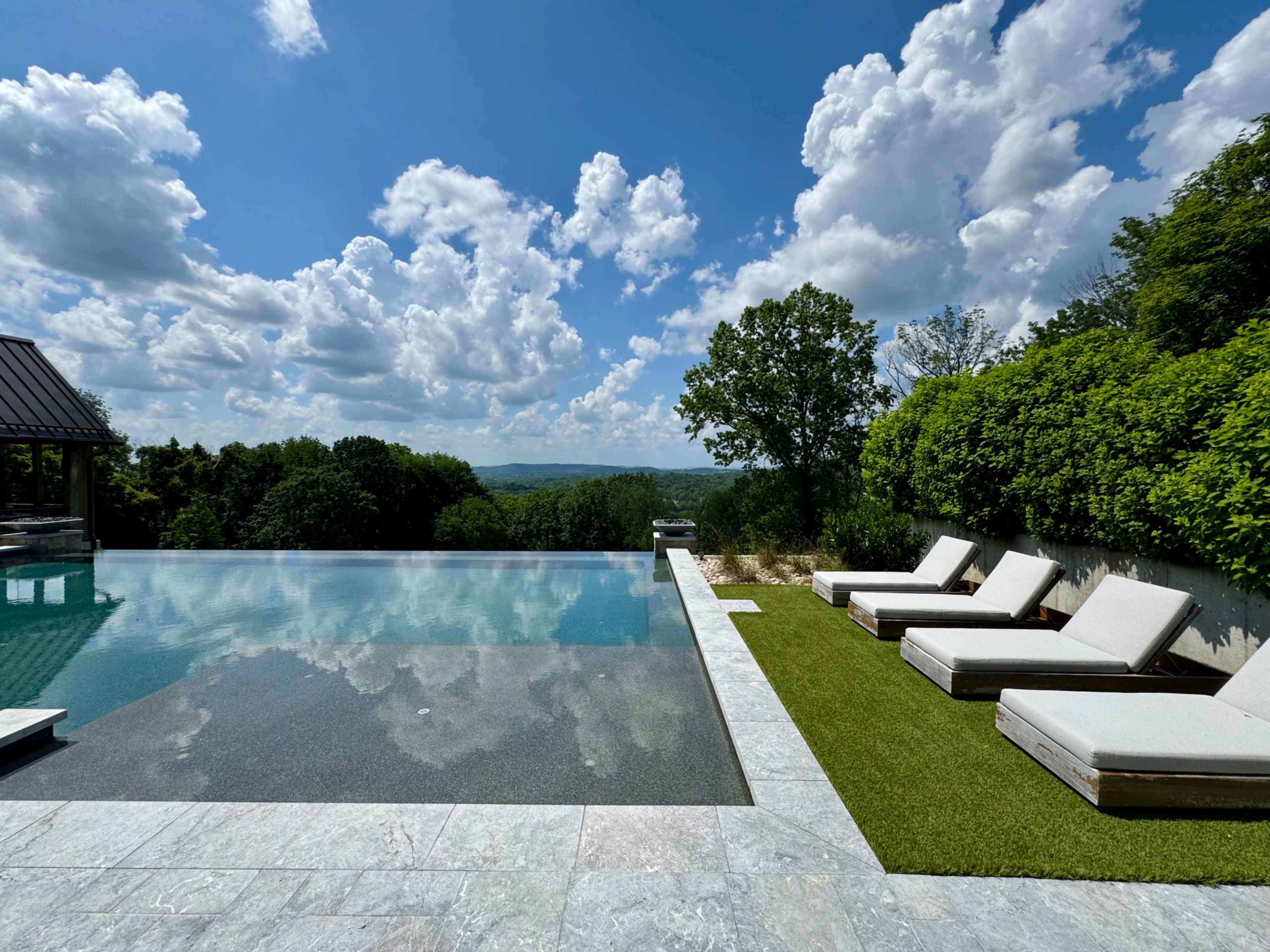 A modern infinity pool with lounge chairs overlooks a lush landscape under a cloudy blue sky.