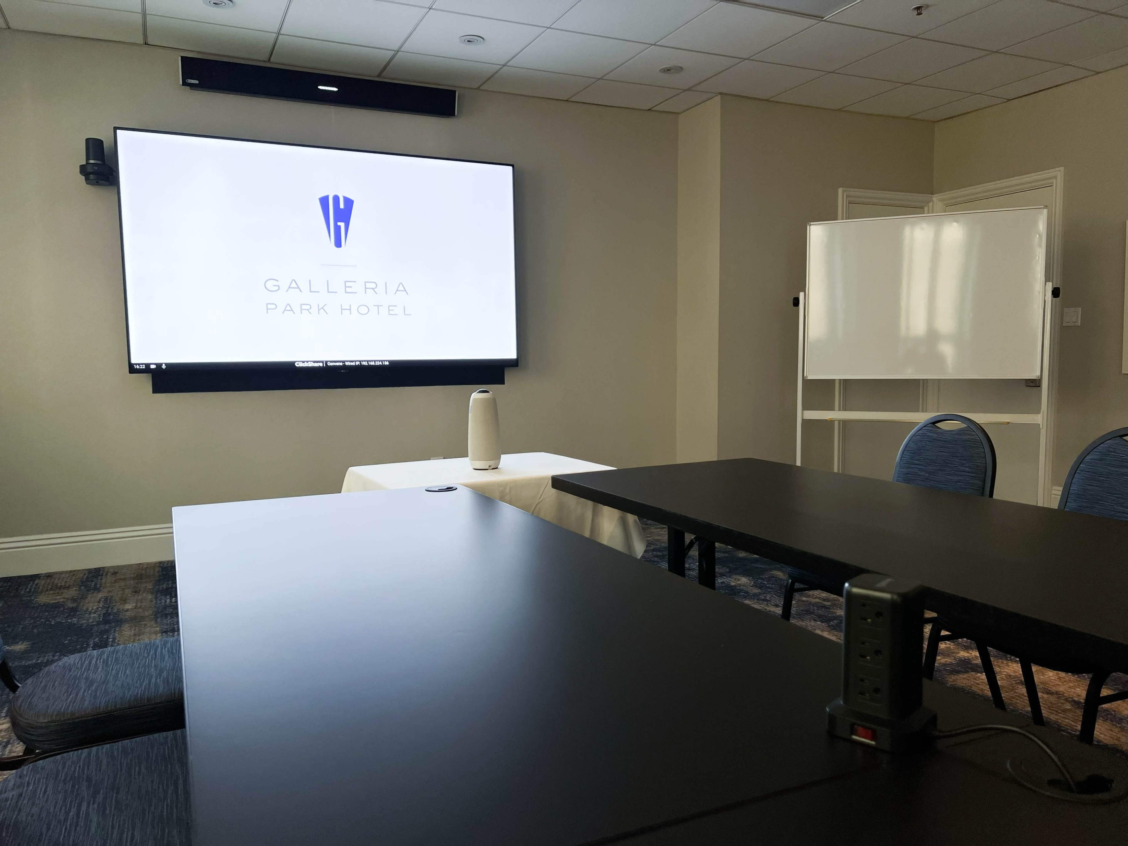 A conference room at the Galleria Park Hotel features a large screen displaying the hotel logo, tables, chairs, and a whiteboard.