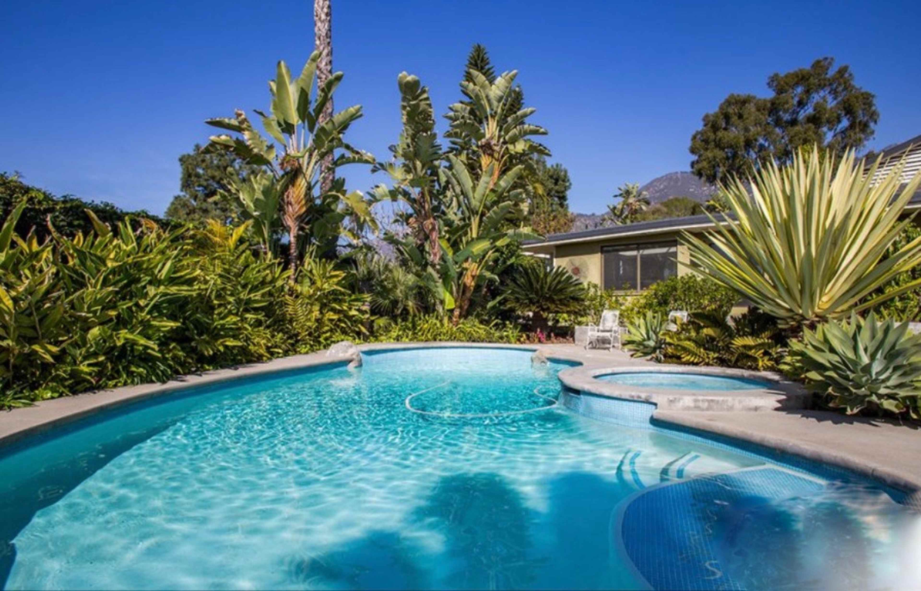 A swimming pool surrounded by lush tropical plants and trees, with a spa area integrated into the pool design.