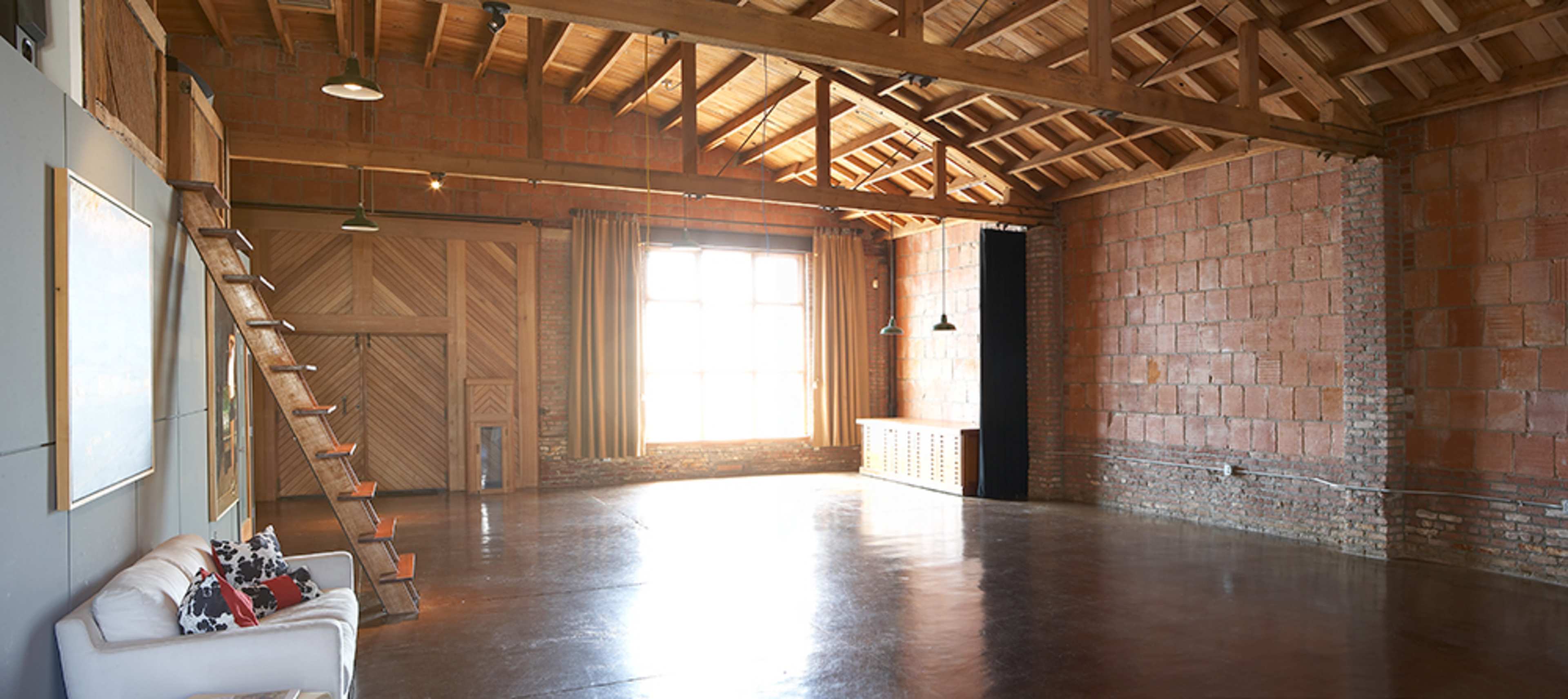A spacious, unfinished interior room with wooden beams, a staircase along one wall, large windows letting in natural light, and a concrete floor.