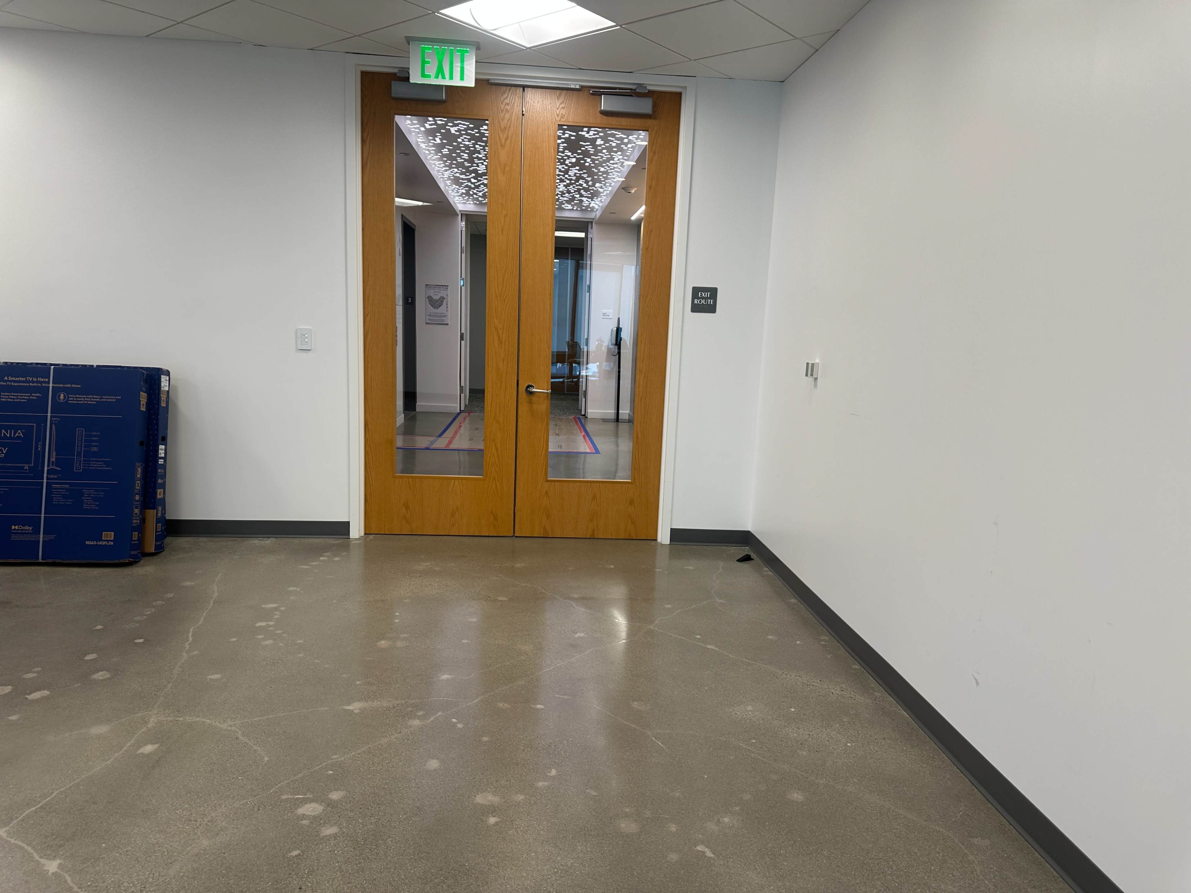 The image shows a pair of wooden double doors leading to an office space, situated next to a blank white wall and a stack of blue boxes on the floor.