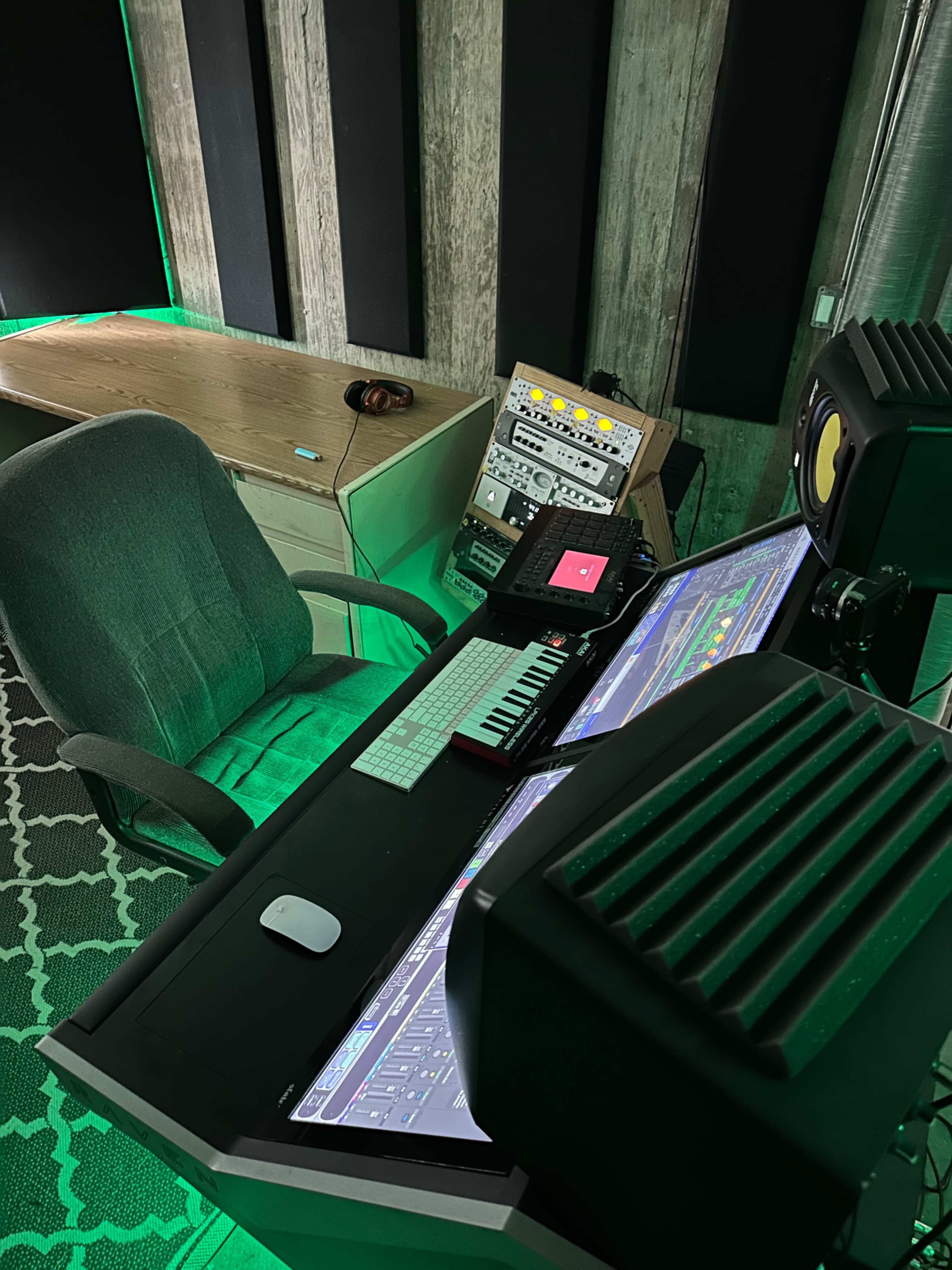 A music production workspace with a computer, MIDI keyboard, and studio monitors set up in a room with green ambient lighting.