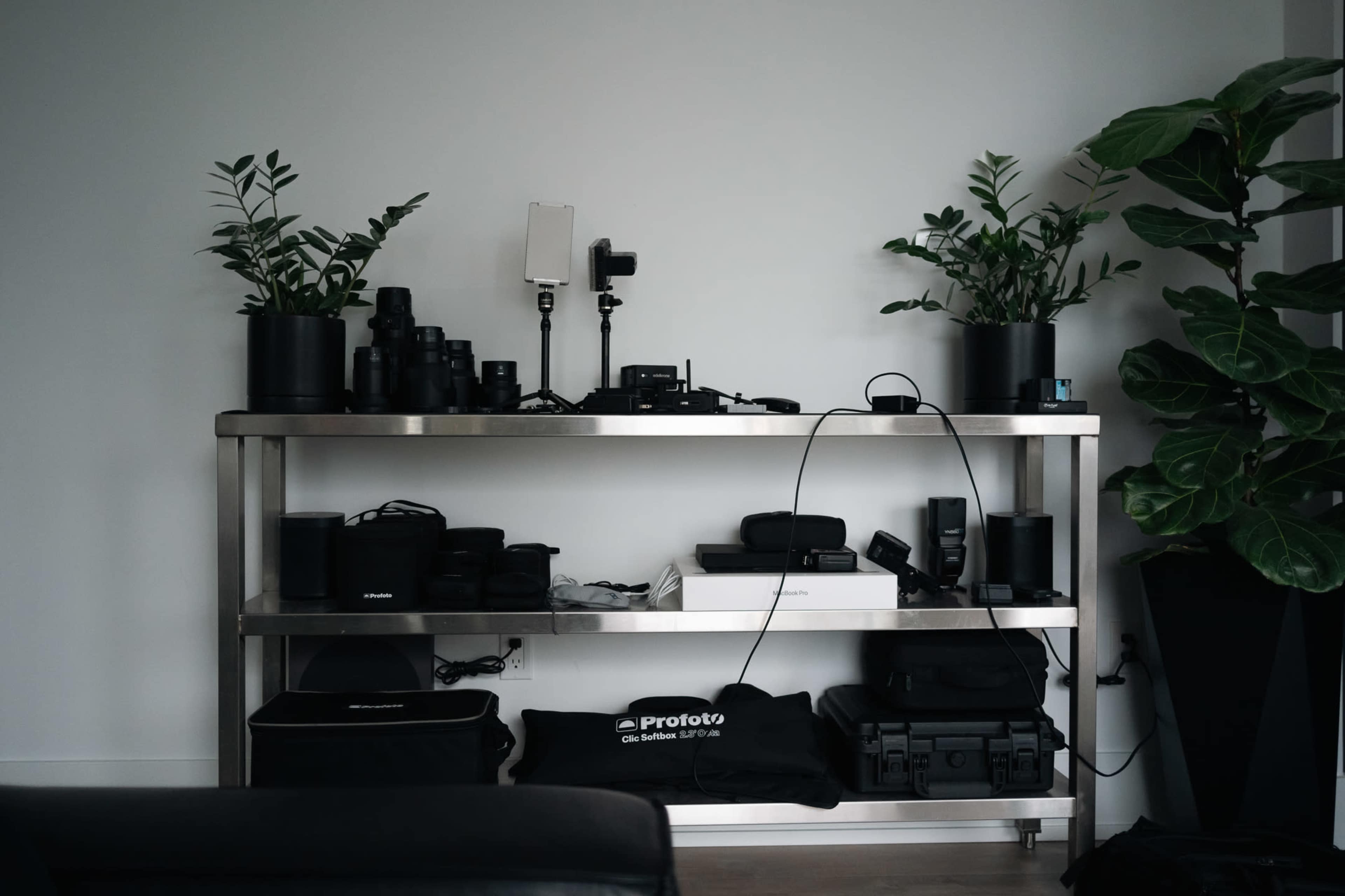 A metal shelf is organized with various photography equipment, including lenses, lights, and plants in pots.
