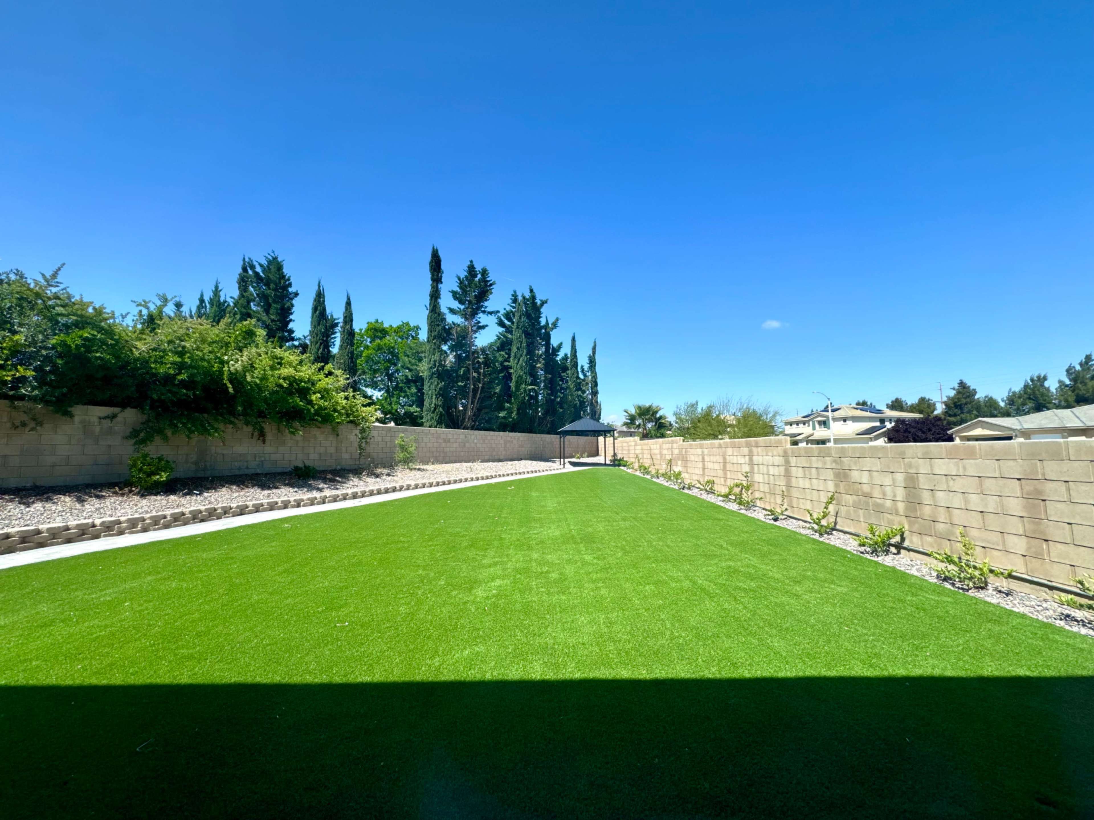 A spacious, well-maintained backyard with artificial grass and a concrete wall surrounded by trees and shrubs.