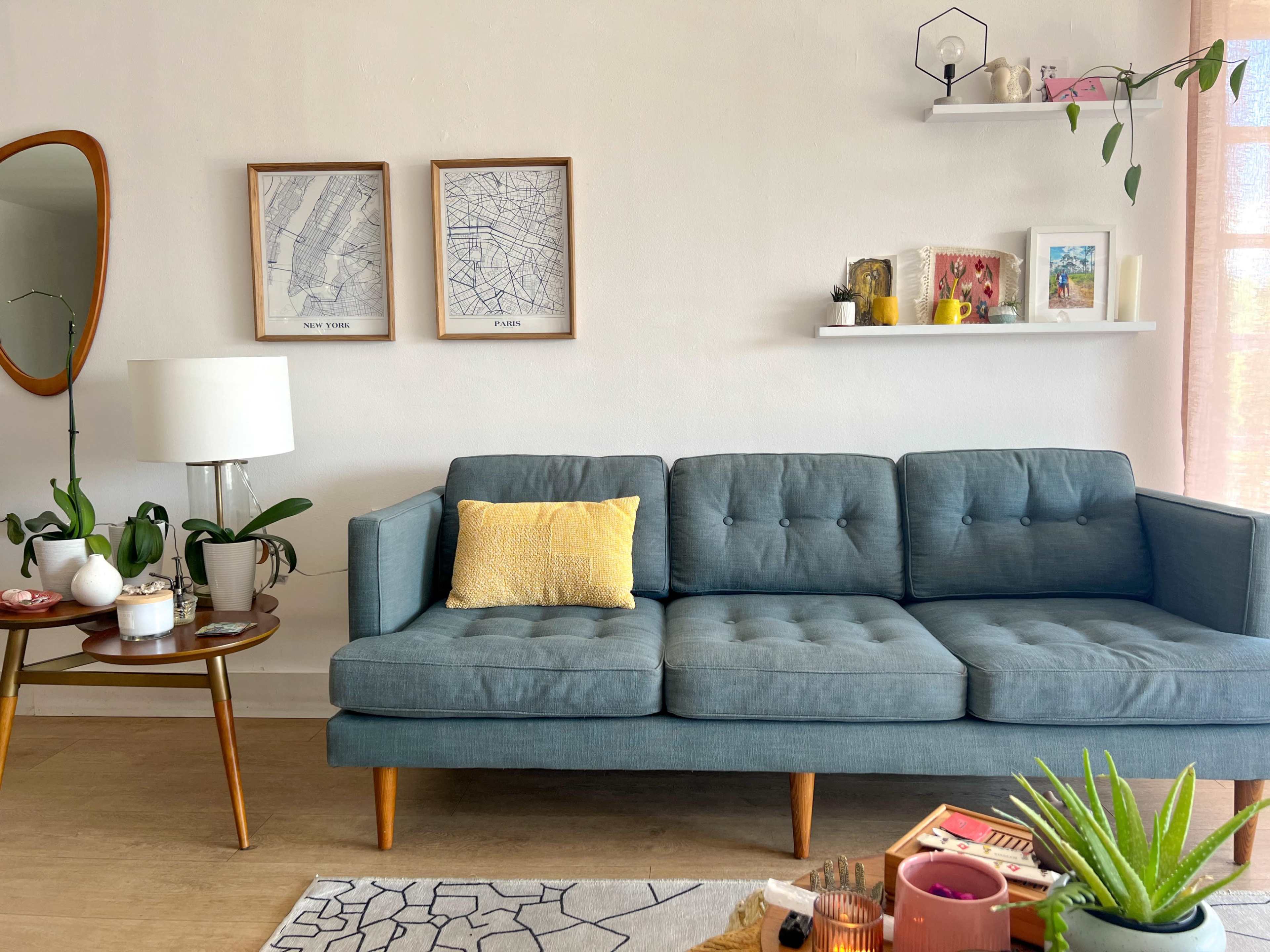 A modern living room features a teal sofa with a yellow cushion, a round side table with plants and a lamp, framed maps on the wall, and shelves displaying decorative items.