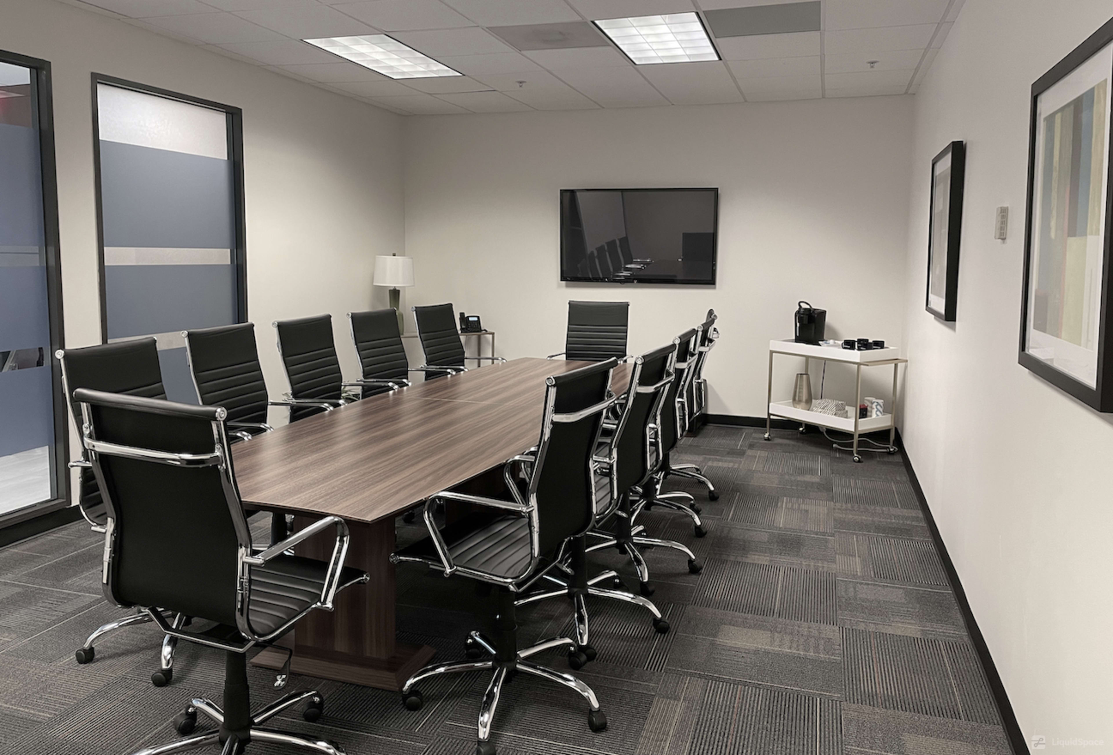 The image shows a conference room with a long wooden table surrounded by black chairs, a television on the wall, and a side console with a coffee maker.